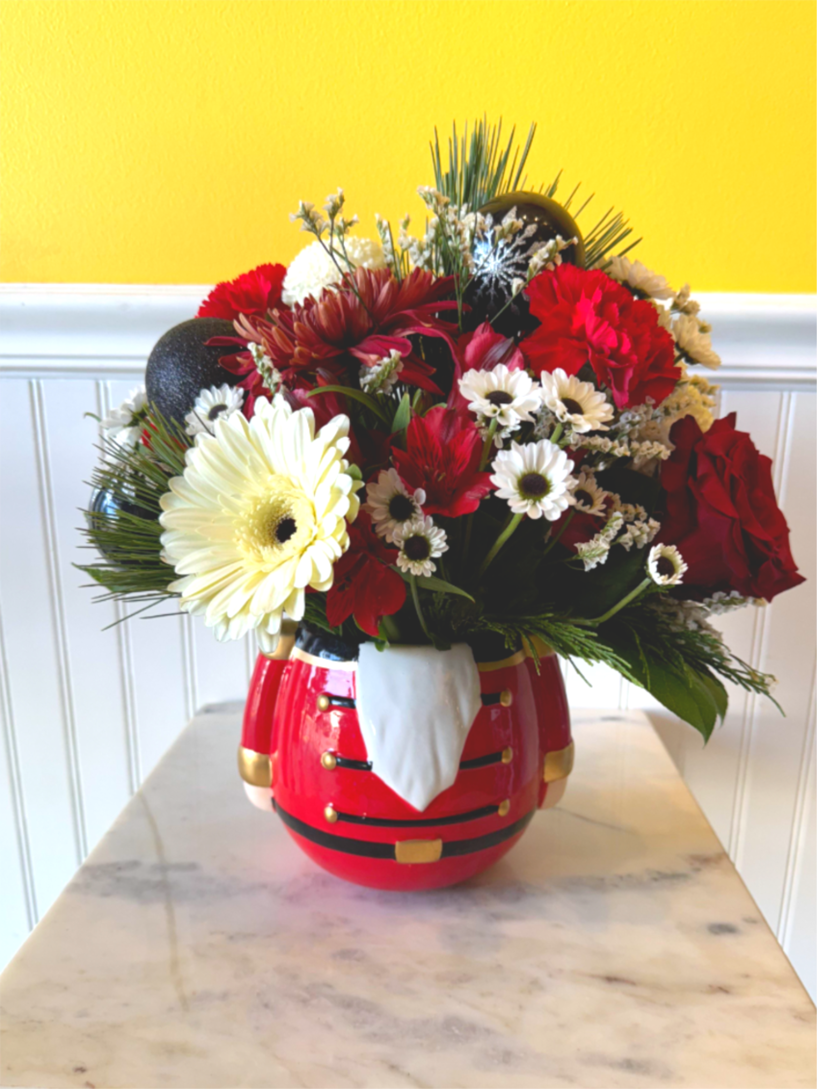 The Festive Nutcracker - Add a dash of holiday charm with The Festive Nutcracker—a cheerful arrangement tucked inside a whimsical toy soldier keepsake. With its bold colors and playful spirit, this piece brings nostalgic joy and seasonal sparkle to any room. Whether you're gifting or decorating, it's a merry little masterpiece that marches straight into hearts. approx Dimensions 14 in X 12 in