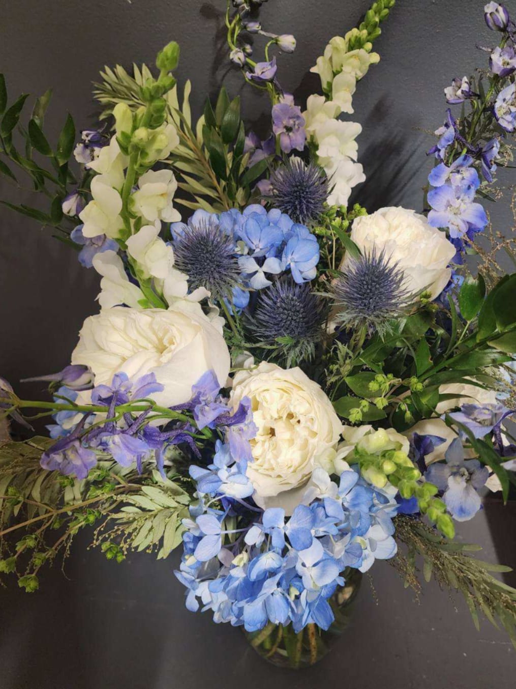 Festival of Lights - A holiday filled with elegance and joy calls for our Festival of Lights centerpiece. Filled with shades of blue, white flowers and lush greenery. Houses in a glass vase.   Height 18-24”  Every effort will be made to recreate your chosen design. Please note that from time to time due to product availability and quality we may need to make substitutions. We will make sure to keep the original design in mind when making substitutions.