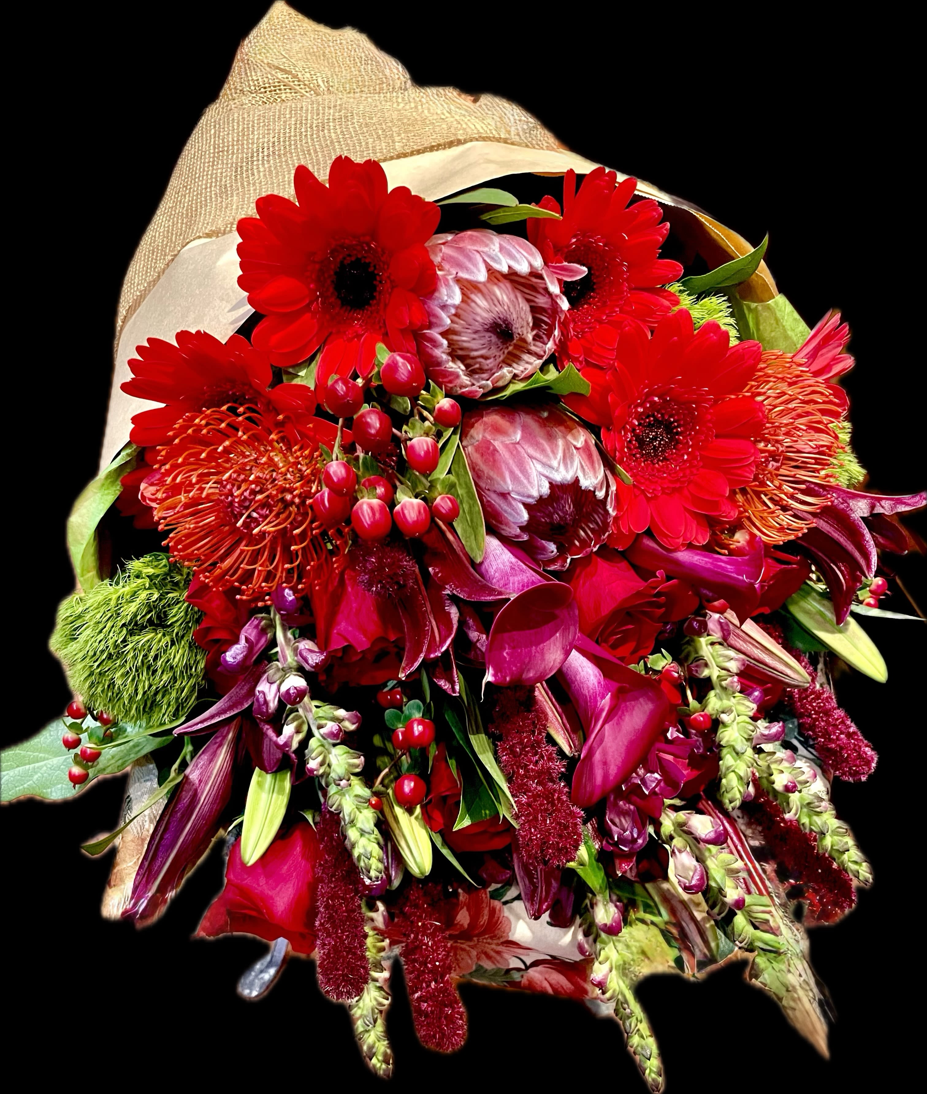 Crimson Noel Bouquet  -  Crimson Noël Bouquet Celebrate the magic of the season with our luxurious Crimson Noël Bouquet — a bold and festive arrangement bursting with rich holiday reds and vibrant textures. This stunning hand-tied bouquet features premium blooms such as striking protea, radiant gerbera daisies, exotic pincushion protea, deep red calla lilies, and elegant roses. Seasonal accents like hypericum berries, lush greens, and cascading amaranthus add depth and a dramatic holiday flair.  Wrapped in natural burlap for a warm, rustic Christmas touch — perfect for gifting, home décor, or as a breathtaking centerpiece for holiday gatherings.   Includes: Premium red florals, seasonal textures &amp; winter greenery  Occasion: Christmas, holiday gifting, festive celebrations  Please note: Flowers may vary slightly based on seasonal availability, while maintaining the same luxurious style and color palette. 