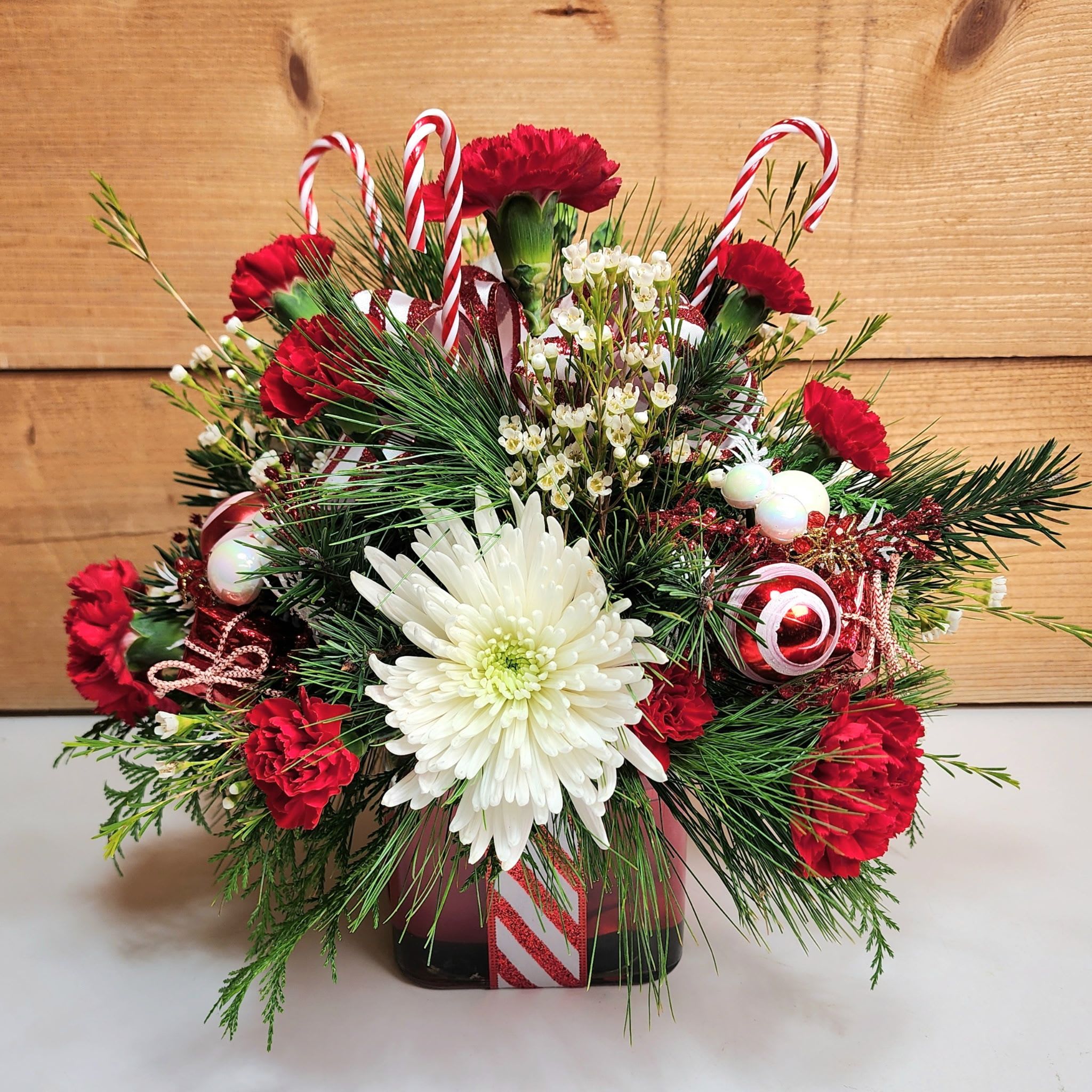 Candy Cane Lane - Candy Cane Lane is a fun arrangement that brings peppermint perfection into your home! Presented in a glass cube with candy cane ribbon and a glittery candy cane bow, this arrangement features red carnations and white mums and waxflower, along with decorative candy cane themed picks for a delightful design that's sure to bring joy this festive season!  Dimensions approx: L 14" x H 15"