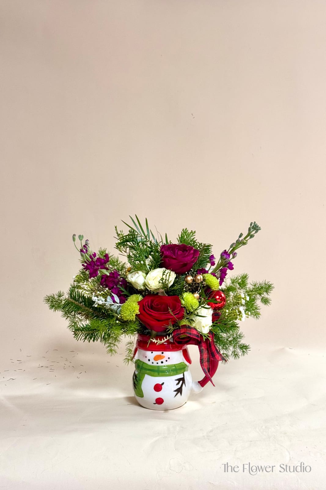 Believe in the Magic - Bring the joy of the season to any space with this festive floral design arranged in an adorable snowman coffee mug. Featuring classic red roses, winter whites, cheerful pops of green, and rich holiday tones, this arrangement is accented with fresh seasonal greens, Christmas ornaments, and a cozy plaid ribbon. Perfect as a gift, a desk display, or a little touch of holiday magic at home, and the mug can be reused all season long! 