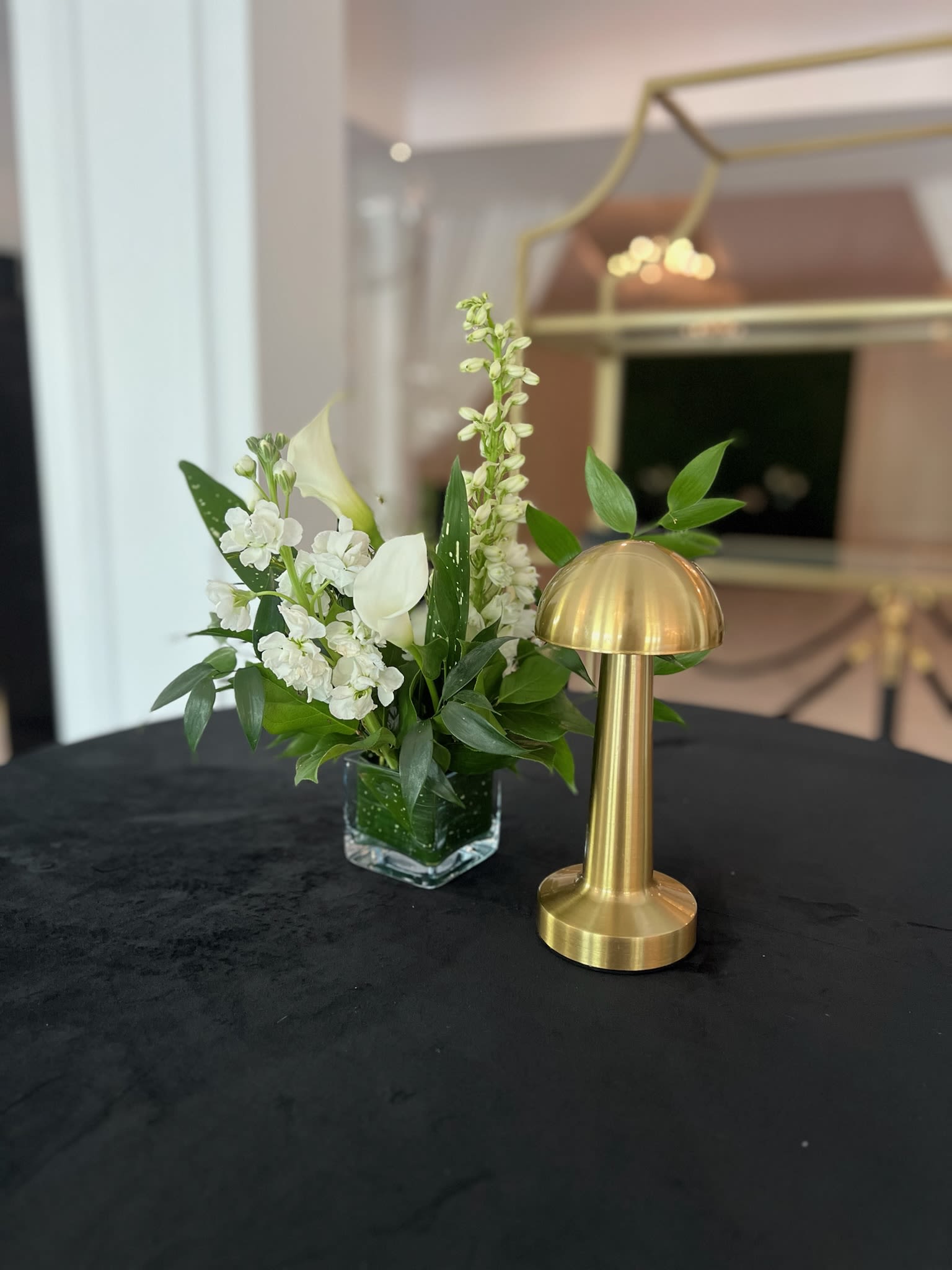 Cocktail Trio Arrangements | Set of 3 - Cocktail Hour Trio Elevate your event with our Cocktail Hour Trio! This set features three chic 3x3 cube arrangements, each bursting with a sophisticated mix of lush greenery and elegant blooms. Choose classic white flowers, or specify up to two colors to perfectly match your theme. These arrangements are ideal for:   * Cocktail tables: Add a touch of refined beauty to your reception.   * Rectangular tables: Group multiple sets together to create a stunning, cohesive centerpiece.  Please note: Gold lamp not included. 