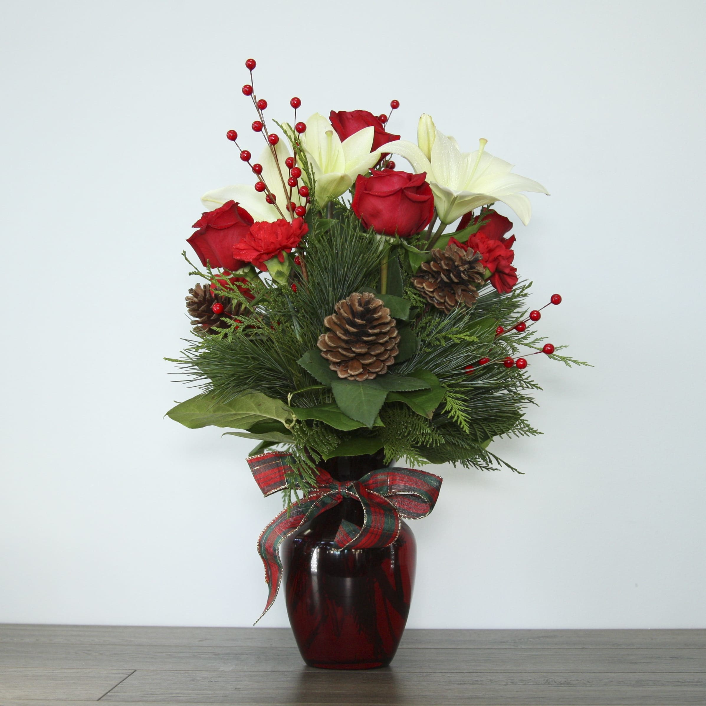 "It's Beginning To Look A Lot Like..." - Centerpiece (EXP 15) - Get in the holiday spirit with this festive centerpiece! This arrangement includes bright red carnations and roses, white Casa Blanca lilies, pine cones, and an assortment of fresh-smelling winter greenery. Minimal Christmas decor and a hand-tied bow is added to complete the look. The centerpiece is placed inside a red tinted glass vase for a classic winter feel. 