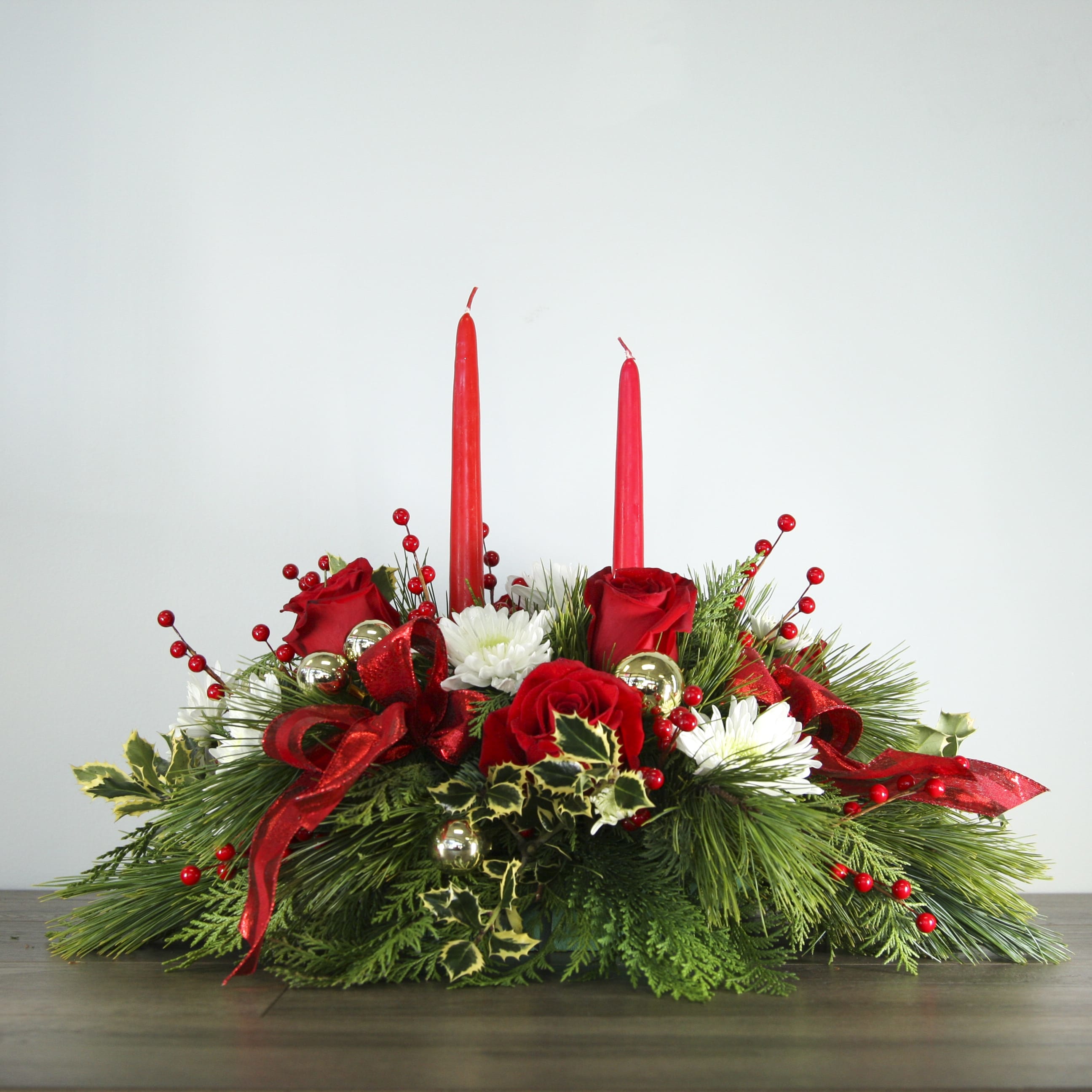 "Let Us Feast!" - Candle Centerpiece (EXP 17) - Gather your friends and family and celebrate around this traditional winter centerpiece! This arrangement features two candles, red roses, white chrysanthemums, holly and an assortment of fresh-smelling winter greenery. Minimal Christmas decor and two hand-tied bows are added to complete the look. 