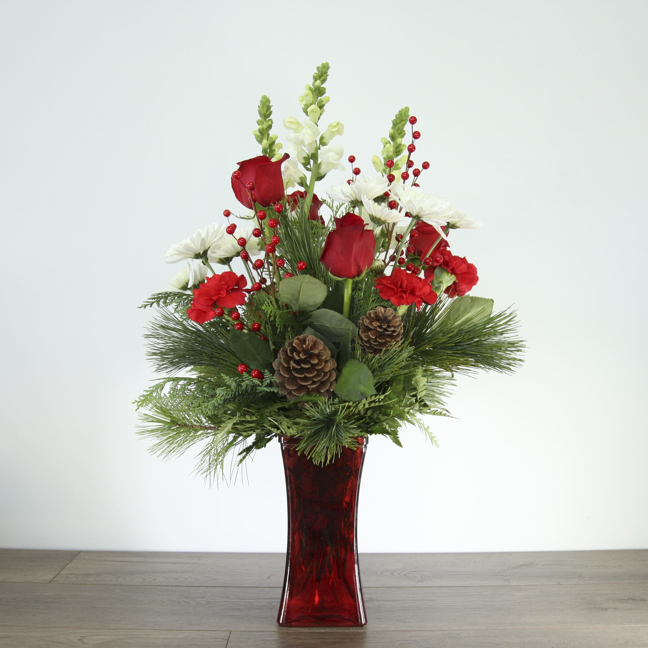Rose Winter Arrangement (EXP 18) - Get in the holiday spirit with this festive centerpiece! This tall and elegant arrangement includes bright red carnations, red roses, white daisies, pine cones, and an assortment of fresh-smelling winter greenery. The centerpiece is placed inside an angular red-tinted glass vase. 