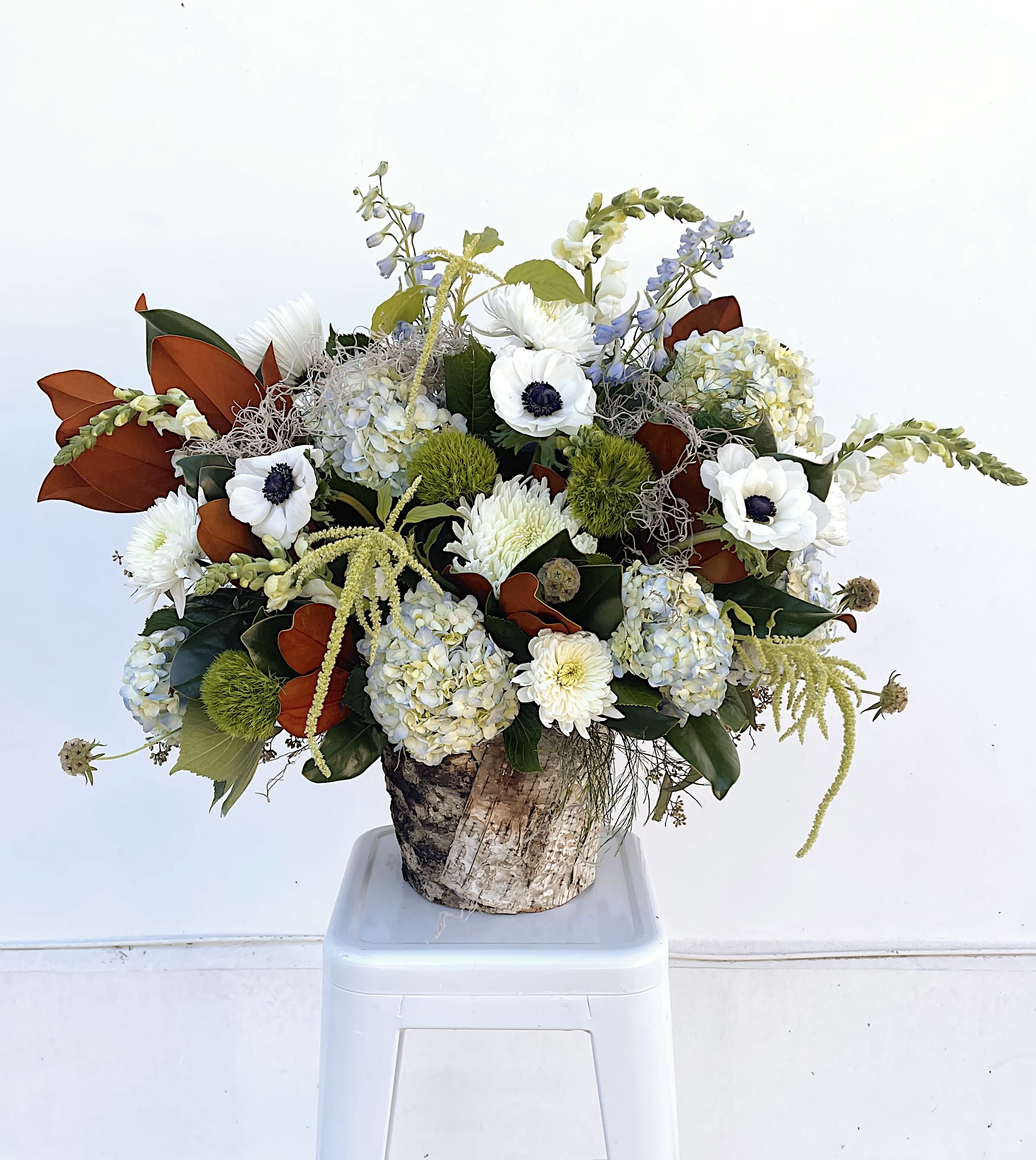 Frosted Elegance - Introducing our new winter flower arrangement, featuring fluffy hydrangeas, lush magnolia leaves, cascading amaranthus, delicate anemones, and elegant snapdragons. This organic, textured blend of seasonal greenery and soft, romantic blooms creates the perfect winter centerpiece to spruce up your holiday parties or your fireside home decor.  ***Smaller size option available by phone***