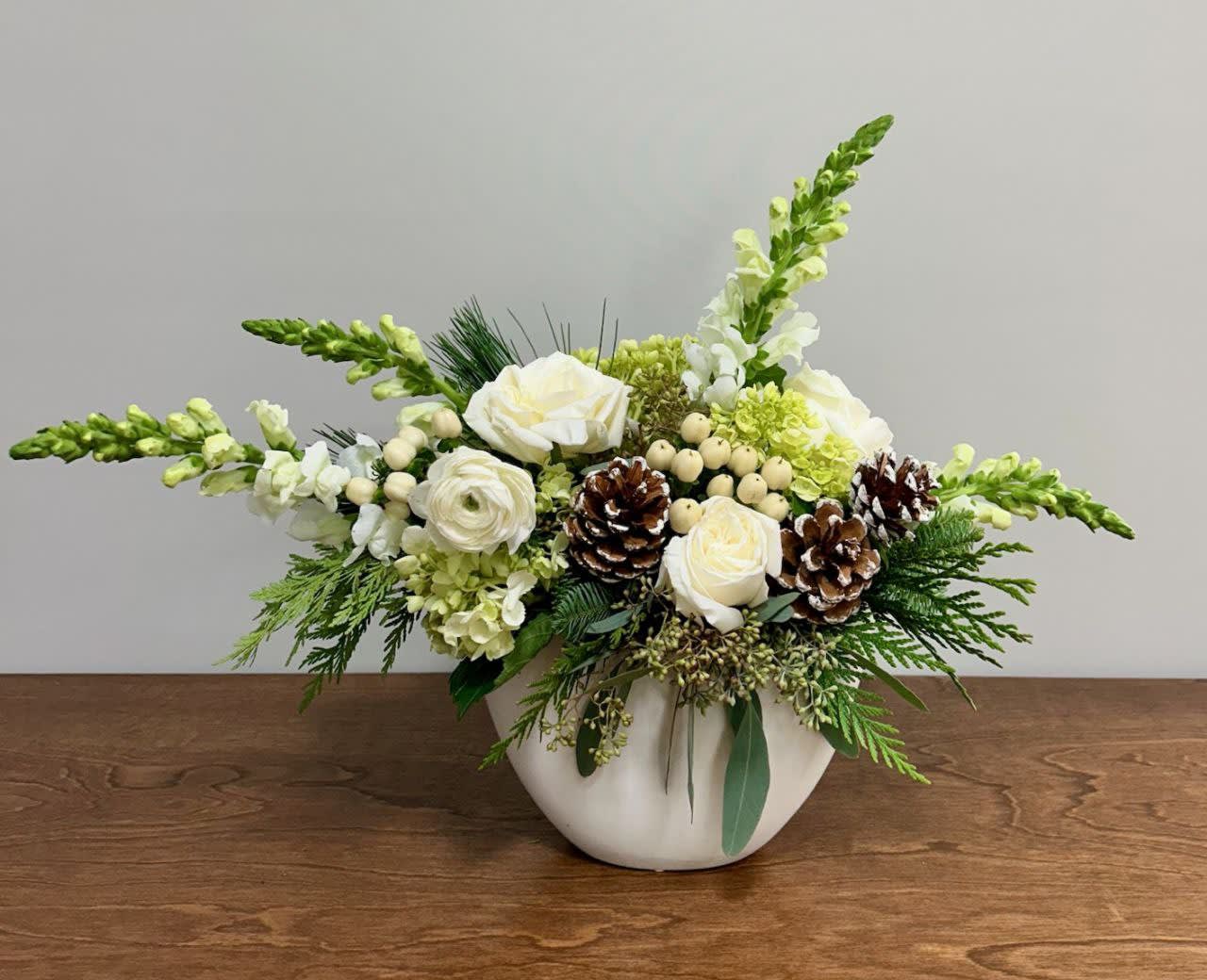 Snowfall - The Snowfall is a harmonious blend of textures, shapes, and hues that transports the viewer to a peaceful winter scene, celebrating the beauty of nature in its quiet and enchanting splendor. White snapdragons, roses, hypericum, ranunculus, mini green hydrangea, and natural pinecones are delicately arranged in a white ceramic oval-shaped vase.