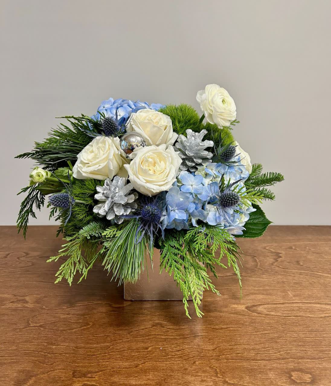Winter Frost - Winter Frost is not just a flower arrangement; it's a captivating symphony of colors, textures, and shapes that brings the enchantment of winter into any space. Perfect for holiday gatherings, winter weddings, or as a thoughtful gift, this arrangement exudes sophistication and timeless beauty. Contains white roses, light blue hydrangea, dianthus, blue thistle, and silver holiday accents in a 5&quot; silver cube.