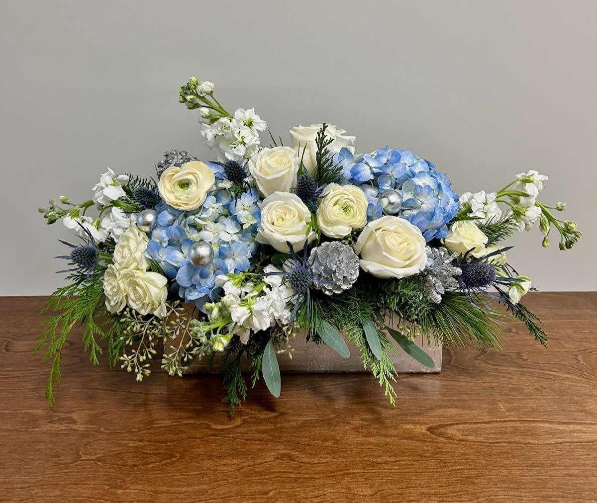 Winter's Nocturne - This wintery flower arrangement features classic white roses, lush blue hydrangea, and festive winter greens, all accented with shimmering pinecones in a long and low silver vase. The winter greens bring a touch of nature indoors while the silver accents add a touch of glamour.