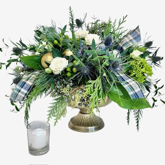 Highland Holiday - This playful arrangement, nestled in a regal gold compote, showcases the spirited hues of blue and green, brought to life by the timeless elegance of tartan ribbon and the proud thistle. Imagine a cozy holiday retreat in the Scottish Highlands, captured in flowers. It's not just an arrangement; it's a merry journey to a land of plaid dreams and festive delight. Embrace the holiday season with a touch of Celtic magic and let the 'Highland Holiday ' transport you to a world of floral joy- with fragrant evergreen, mini green hydrangea, white spray roses, and green hypericum berries. Added holiday flair from golden baubles. 