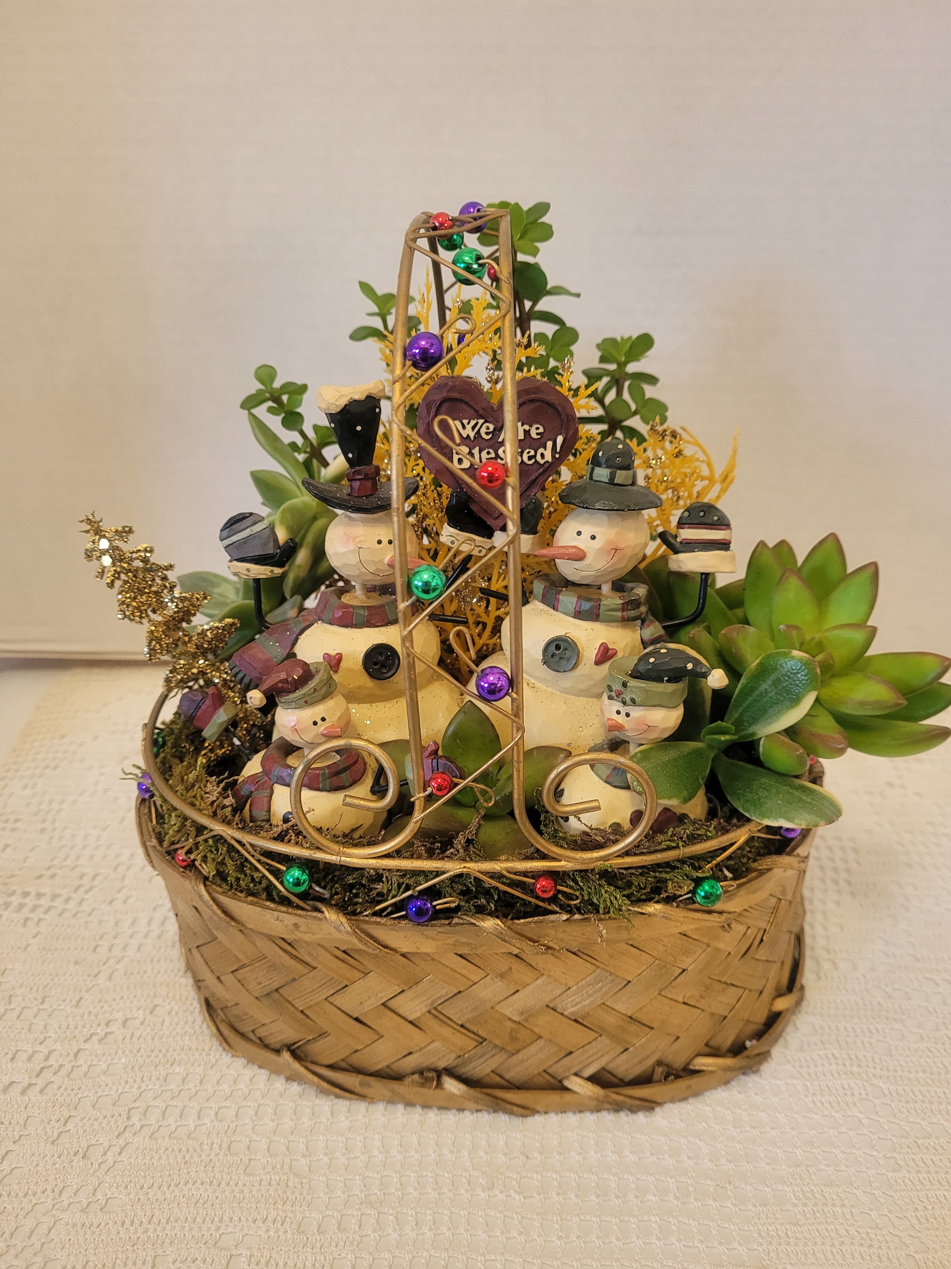 We Are Blessed - This wire and woven basket is filled to the brim with succulents but the charm of this arrangement is the ceramic snowman family settled inside. They are ready to celebrate with you.