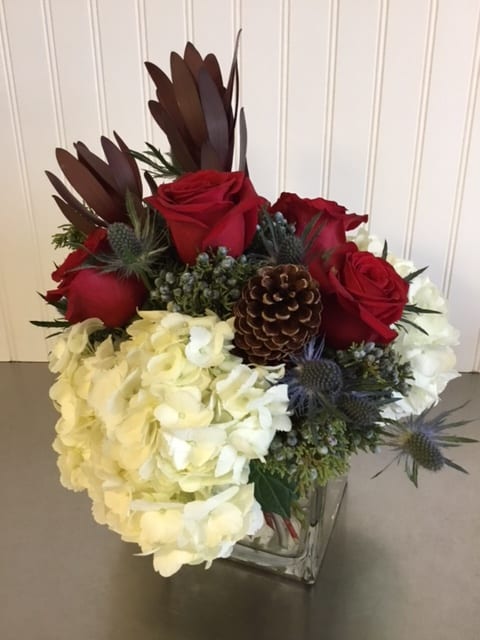 Adeline - For the love of the season, a snowy collection of white Hydrangea and steel colored Thistle complemented with red Roses and tall Protea. The arrangement is supported with greens and festive Pinecones.