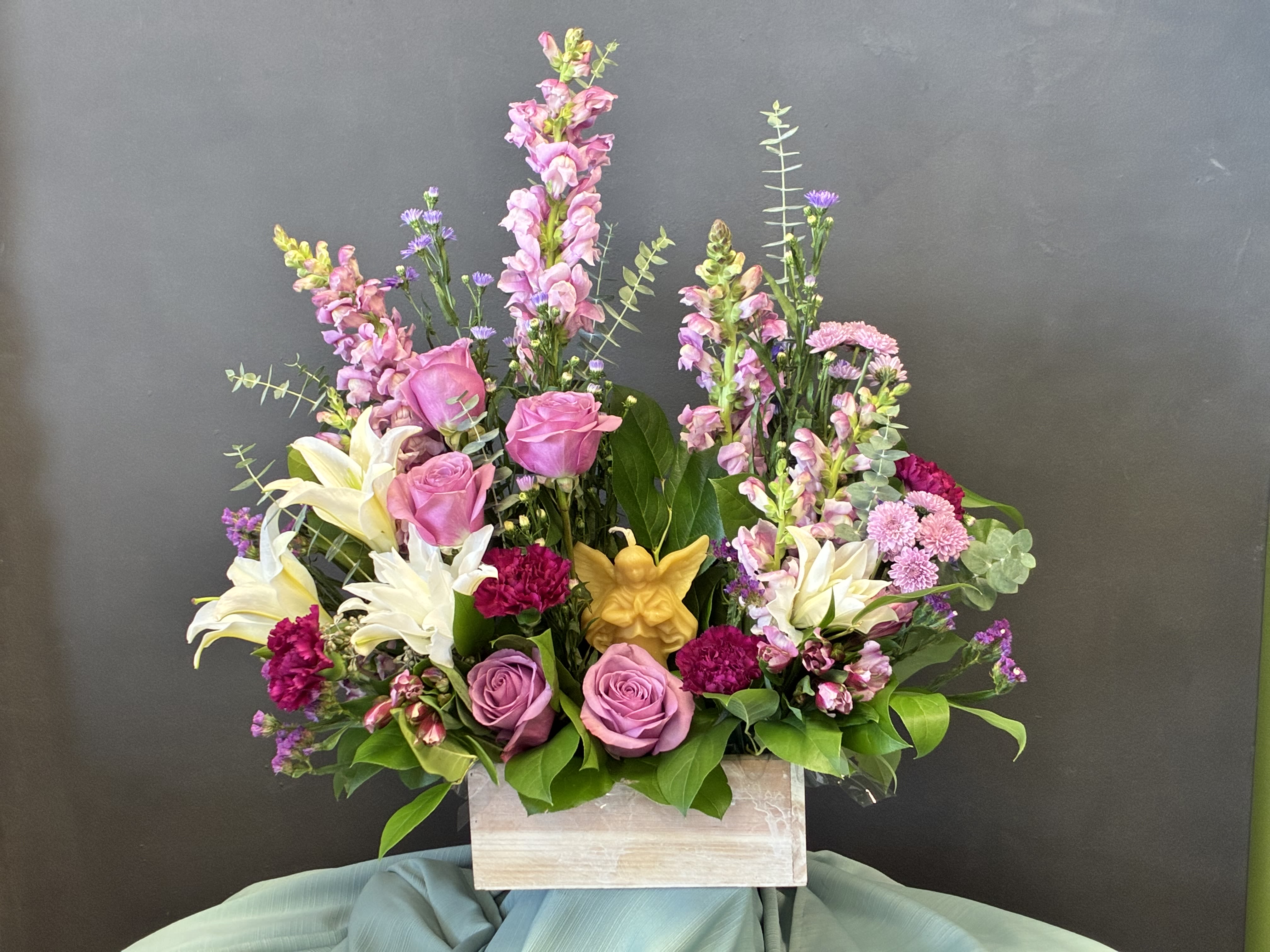  Heavenly Holiday Grace - Celebrate the spirit of Winter with this heartwarming arrangement, nestled in a rustic wooden box and crowned with a golden angel candle —a symbol of peace, hope, and divine love. Blush roses and creamy lilies evoke winter’s softness, while rich burgundy carnations and violet snapdragons add festive depth. Sprigs of lavender and pastel filler blooms mingle with lush greenery, reminiscent of frosted pine and holiday garlands. Perfect for a mantelpiece, memorial, or holiday centerpiece, this bouquet brings comfort and beauty to the season of giving.