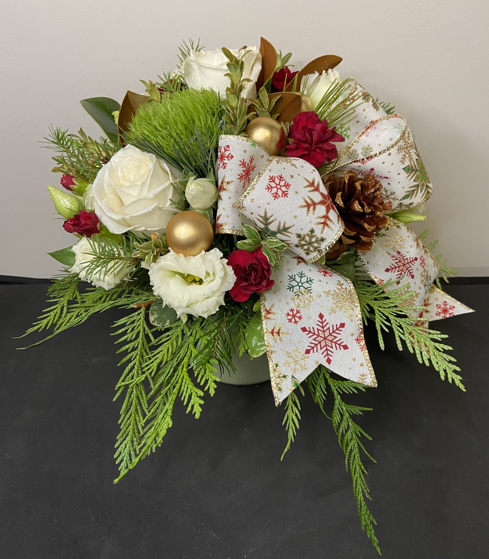 North Pole Noel by Chasing Petals - Elevate your holiday décor with our “North Pole Noel” arrangement. This sophisticated design features creamy white blooms, rich red accents, and luxurious evergreens, complemented by gold ornaments and a delicately patterned snowflake ribbon. Finished with natural pinecones for a touch of winter texture, this arrangement offers a graceful blend of seasonal elegance and timeless festive charm. Perfect as a holiday centerpiece or a refined seasonal gift.
