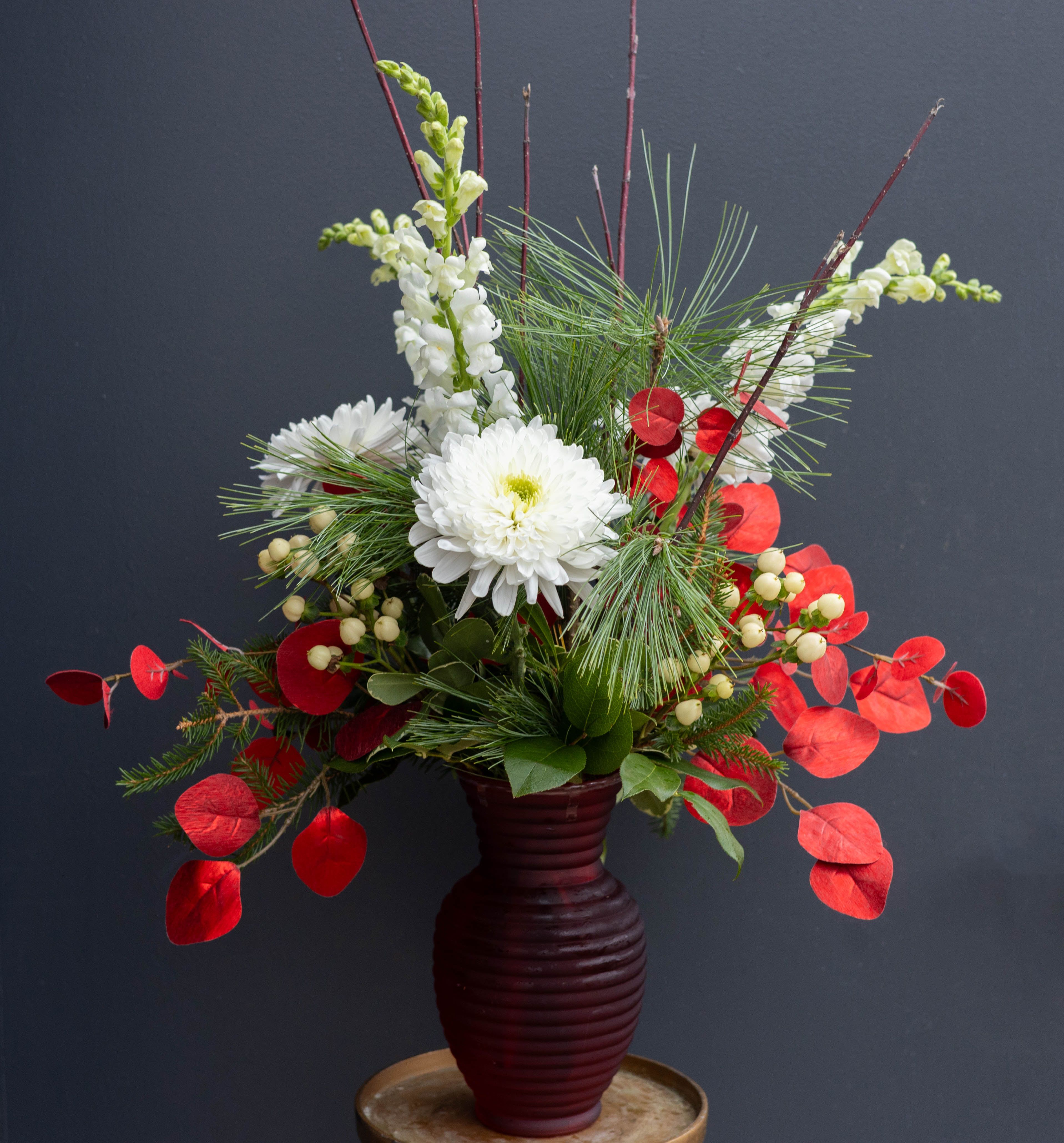 Holiday Blitz - Real &amp; Faux Flower Mix - Bursting with festive energy, Holiday Blitz is a striking Christmas arrangement that blends classic holiday colors with modern seasonal texture. Crisp white blooms rise gracefully among tall winter greens, creating a fresh, snowy contrast. Vibrant red accents—featured through bold, round foliage—add a cheerful pop of color that instantly captures the spirit of the season. Soft white berries and long, airy pine needles bring depth and movement, while slender red branches give the design an elegant vertical lift.  Presented in a rich, deep-red vase, this arrangement feels both warm and sophisticated. Holiday Blitz is perfect for brightening an entryway, adorning a holiday table, or gifting as a joyful celebration of winter’s natural beauty.