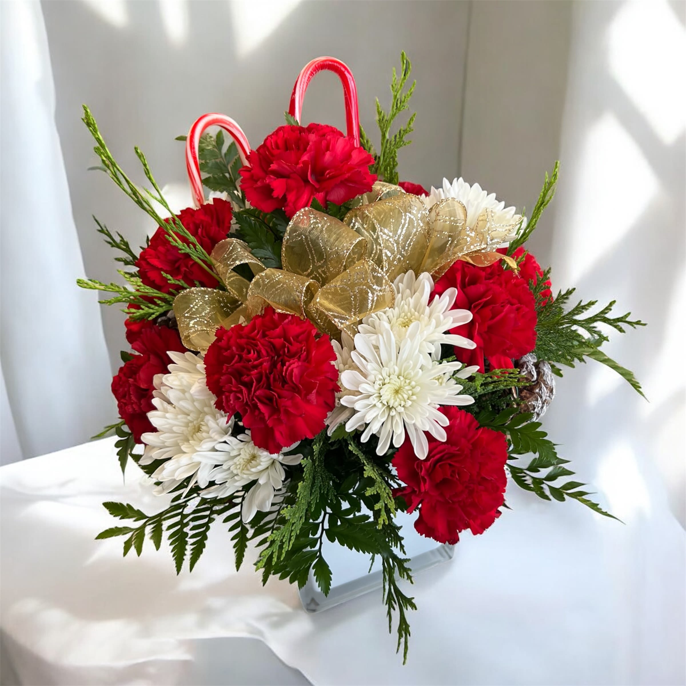 Sweet Mint Joy  - Sweet Mint Joy is a festive Christmas arrangement designed to brighten any space with holiday cheer. It features vibrant red carnations paired with elegant white spider mums, complemented by fresh winter greenery for a lush, seasonal look. Candy canes add a playful, traditional touch, while the gold ribbon brings a hint of sparkle and sophistication. Perfect for gifting, decorating your home, or adding a joyful accent to any holiday celebration. A delightful blend of sweetness, elegance, and Christmas magic!  Sweet Mint Joy es un arreglo navideño festivo diseñado para iluminar cualquier espacio con el espíritu de la temporada. Incluye vibrantes claveles rojos combinados con elegantes flores blancas tipo spider mum, acompañados de follaje fresco que aporta un toque invernal. Los bastones de caramelo añaden un detalle dulce y tradicional, mientras que el listón dorado aporta un brillo elegante y sofisticado. Perfecto para regalar, decorar tu hogar o añadir un toque alegre a cualquier celebración navideña. Una mezcla encantadora de dulzura, elegancia y magia navideña!