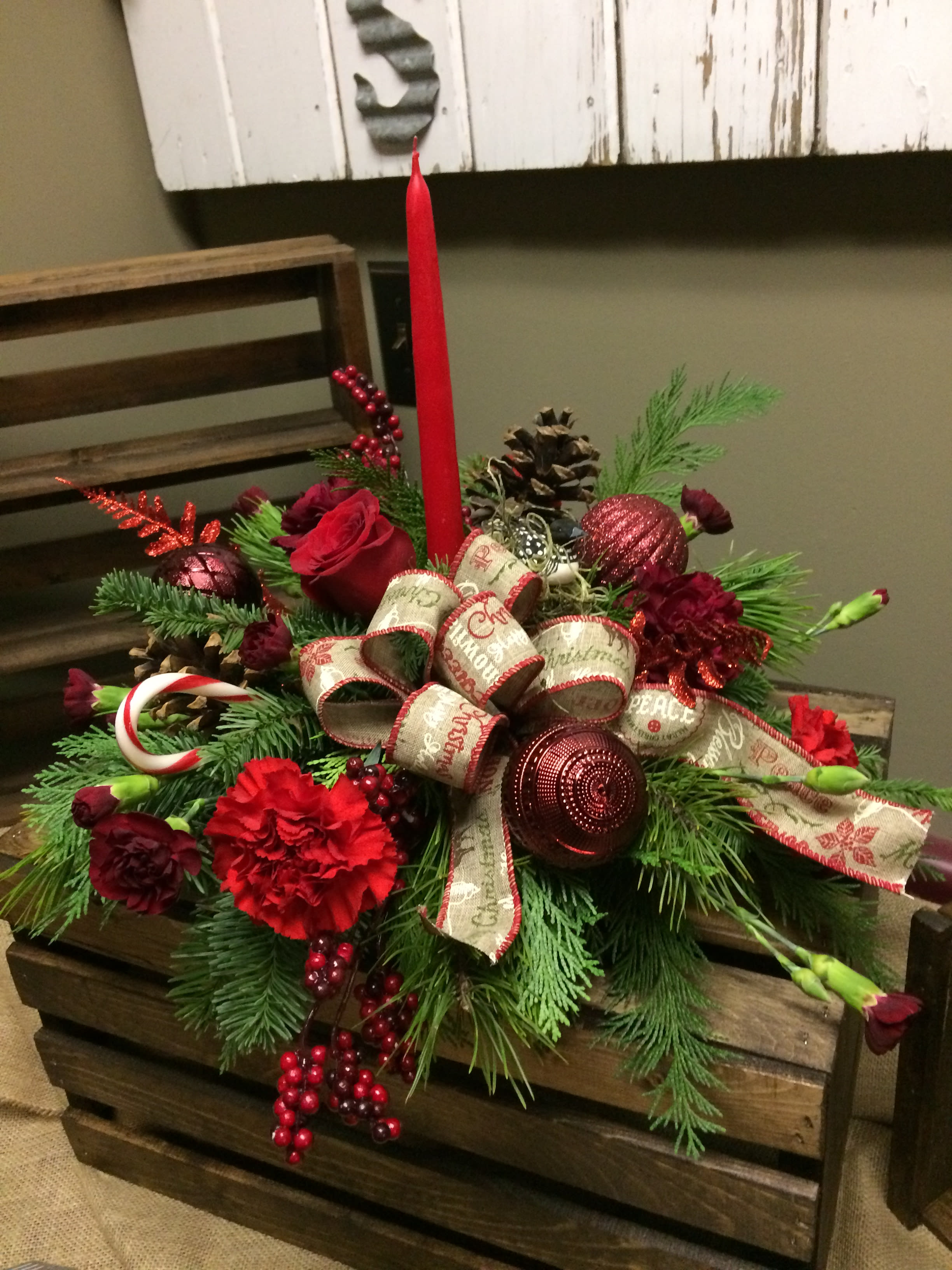 Seasons Bright Centerpiece Custom Design by Cold Spring Floral - Seasons Bright Centerpiece is expertly designed in an economical design dish with red and burgundy standard carnations, burgundy miniature carnations, a variety of fresh pine boughs and pine cones. Nestled in the center a red roses and a red taper candle. Accented with  holiday bulbs, artificial red berries and real candy canes; Seasons bright is sure to add lasting Holiday Spirit to any space. Exact colors of flowers and accents are subject to change based on  availability and may vary. This centerpiece is available in other color variations, give us a call today to order!