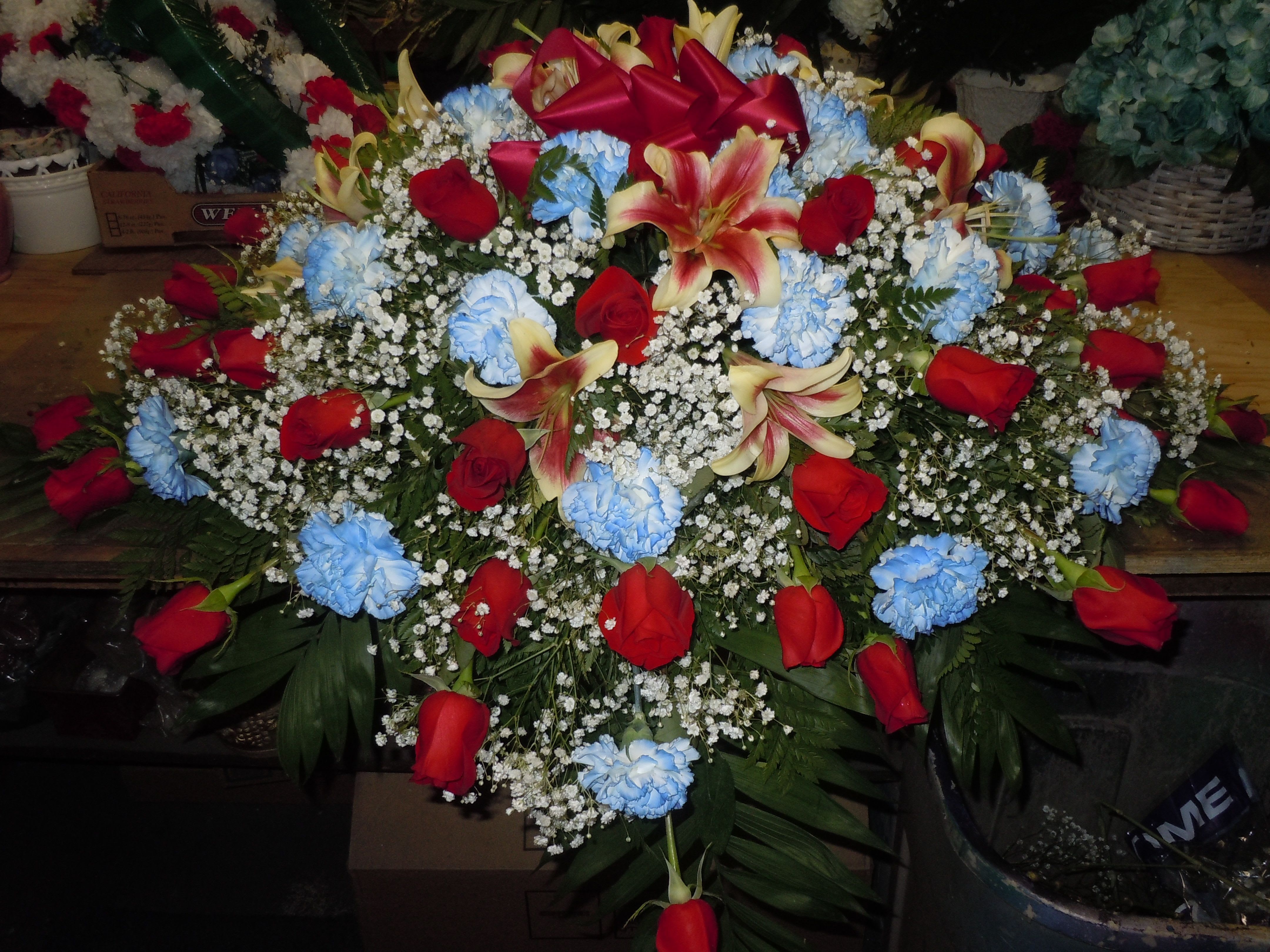 C14 Casket Spray - A mixture of white carnations tinted delphinium blue, red roses, shocking lilies, greens, and baby's breath.   Banner - $15.00 + extra  Substitutions may apply, pending flower availability.