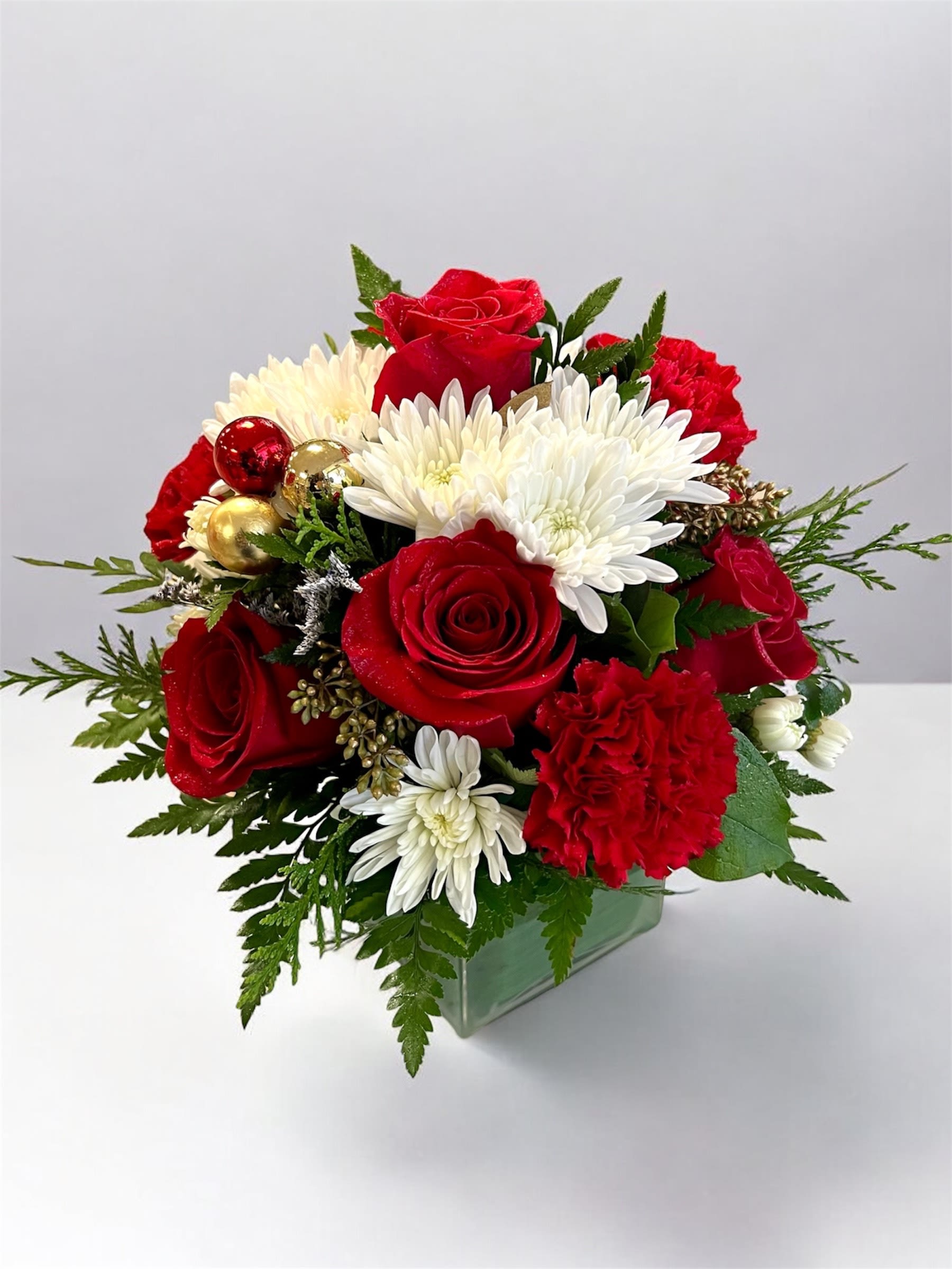 Holiday Sparkle  - The Holiday Sparkle Bouquet is a festive blend of classic Christmas colors and elegant seasonal textures! Featuring vibrant red roses, red carnations, and crisp white chrysanthemums, this arrangement brings warmth and joy to any holiday space. Accented with golden and red ornaments, winter greenery, and arranged in a modern glass cube, it’s the perfect centerpiece to brighten celebrations or share as a thoughtful holiday gift.  El Ramo Brillo Navideño combina los colores clásicos de la Navidad con texturas elegantes de la temporada! Con rosas rojas, claveles rojos y crisantemos blancos, este arreglo aporta calidez y alegría a cualquier espacio festivo. Decorado con esferas doradas y rojas, follaje invernal y presentado en un cubo de vidrio moderno, es el centro de mesa perfecto para iluminar las celebraciones o regalar en estas fiestas.