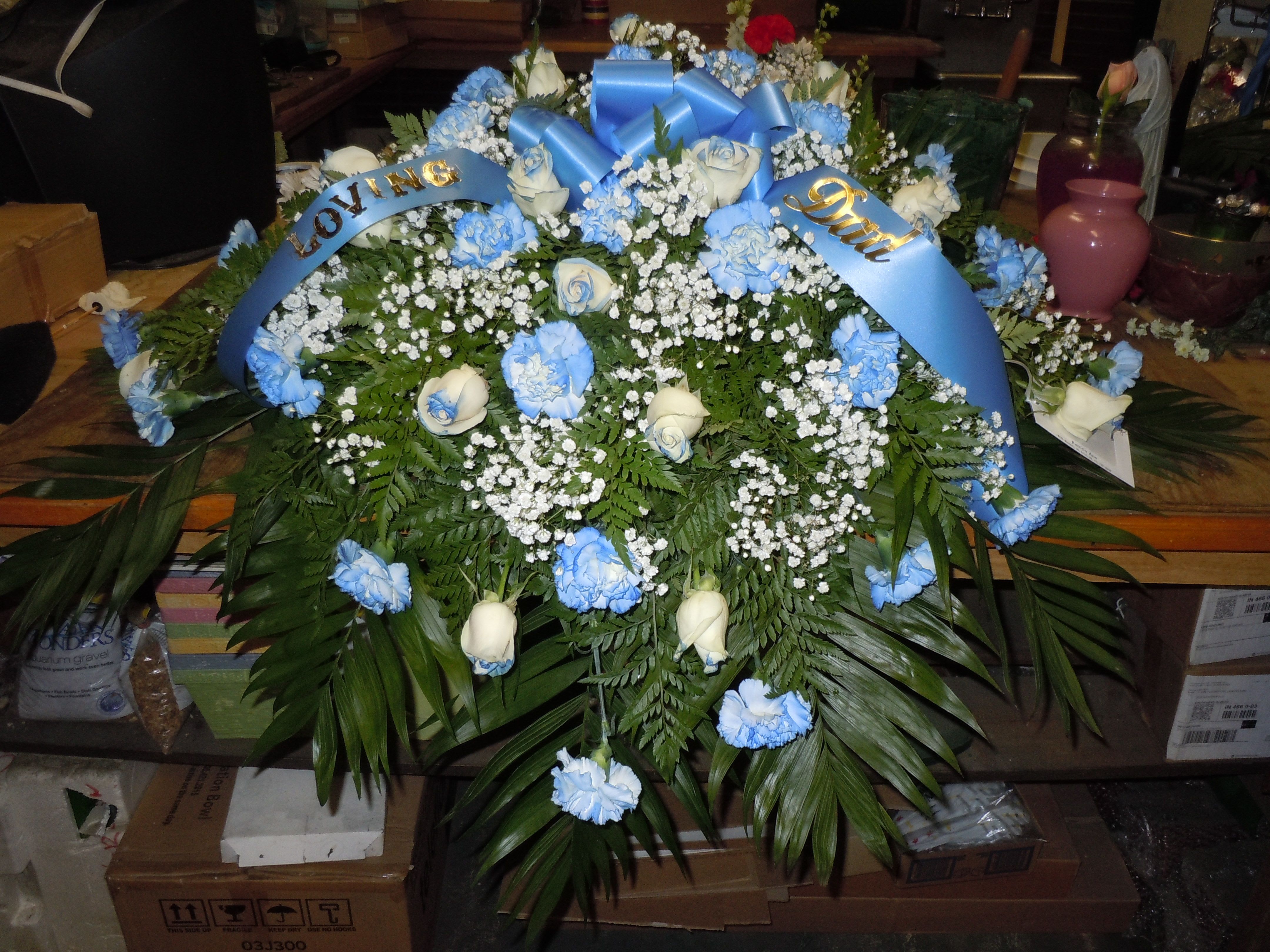 C22 Casket Spray - A mixture of white carnations in a medium blue shade, vendela roses tipped medium blue, greens, and baby's breath, includes bow.  Banners are $15.00 +   Substitutions may apply, pending flower availability.