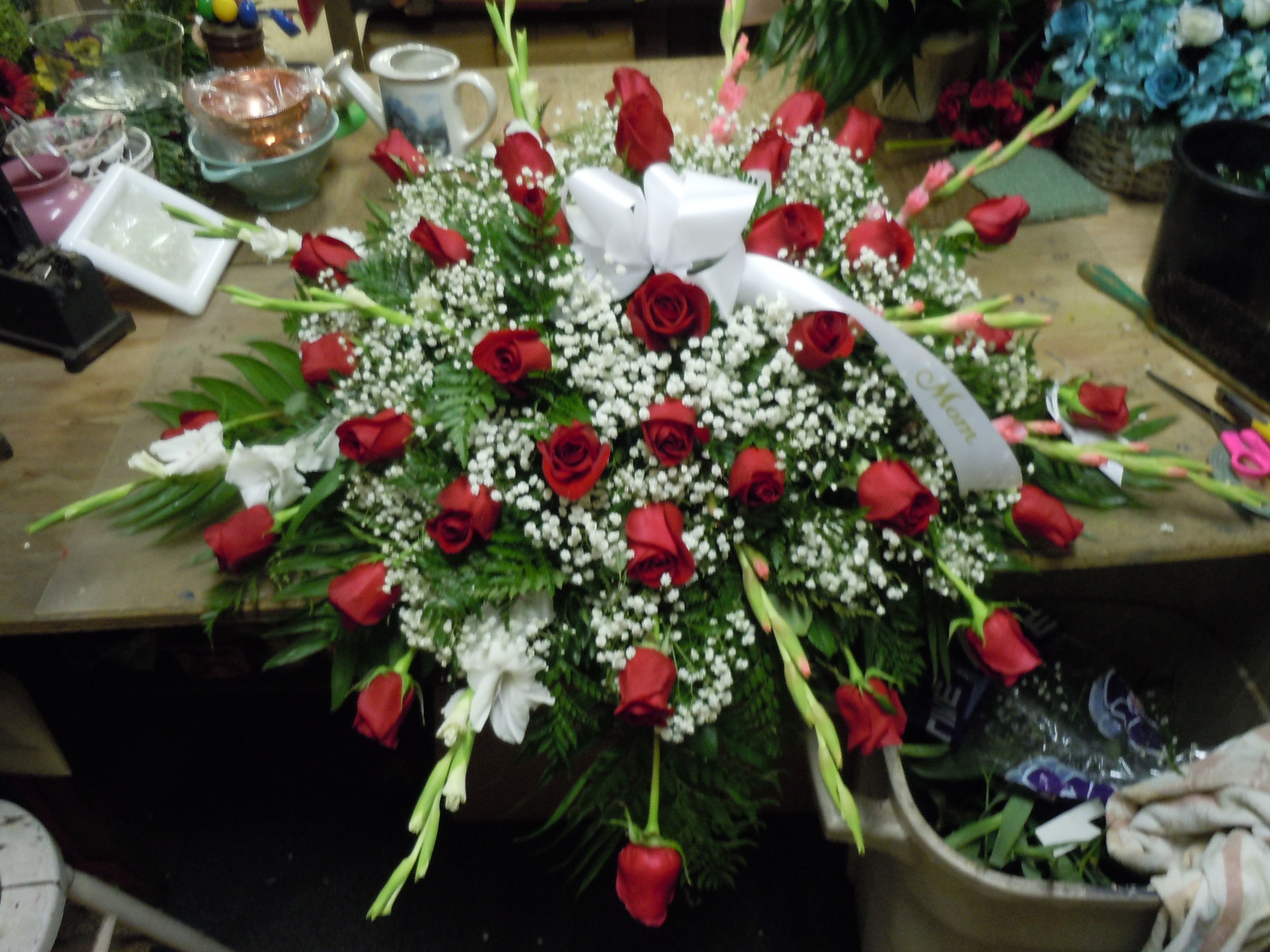 C23 Casket Spray - A mixture of roses, white and red gladiolas (will sub if unavailable), greens, and baby's breath, includes bow.  Banners are $15.00 +   Substitutions may apply, pending flower availability.