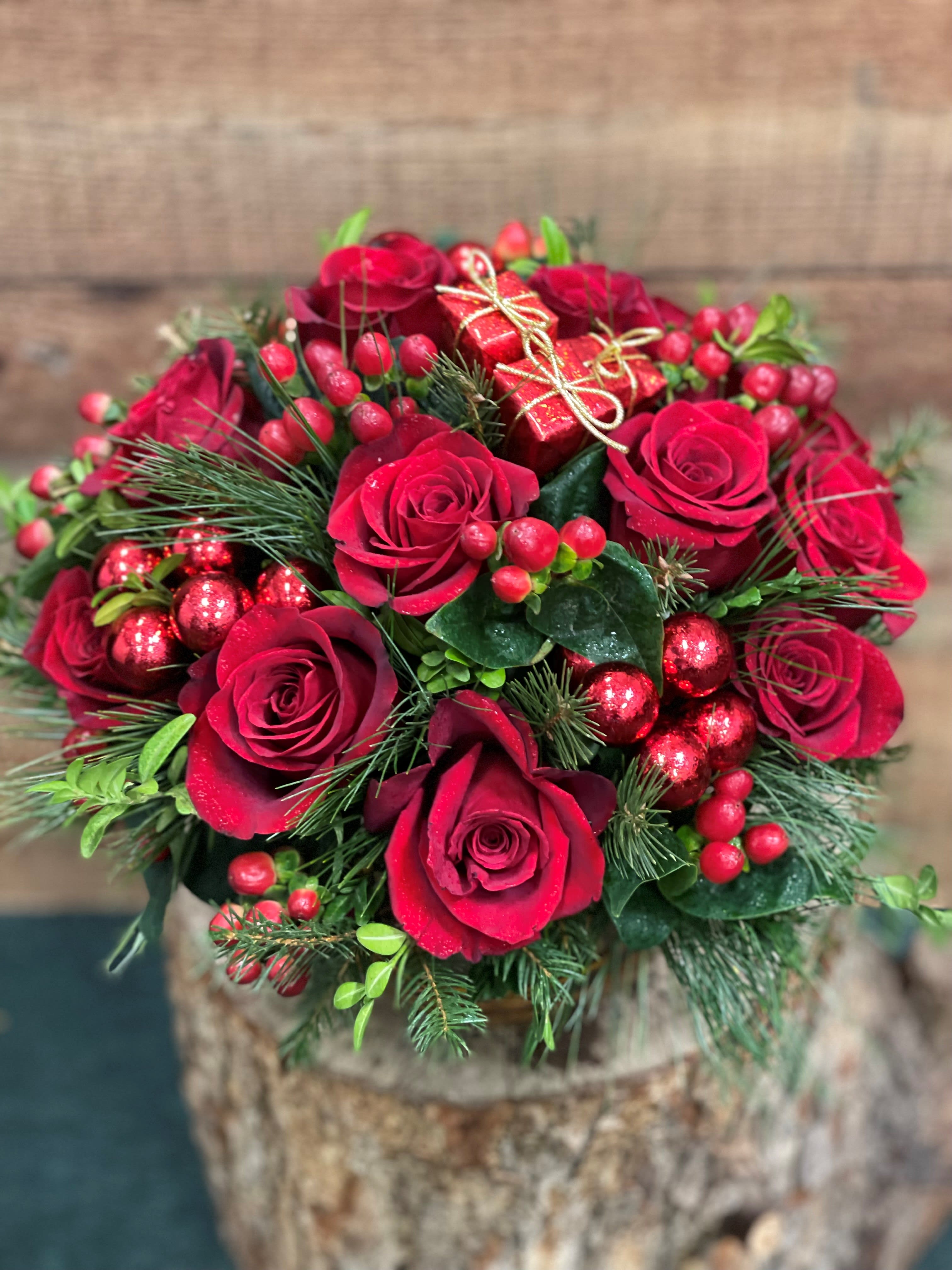 Christmas Rose Vase - Vibrant Red Roses are accented with holiday greens and red hypericum berries in a low vase. With just a touch of added sparkle with glass balls and some packages.