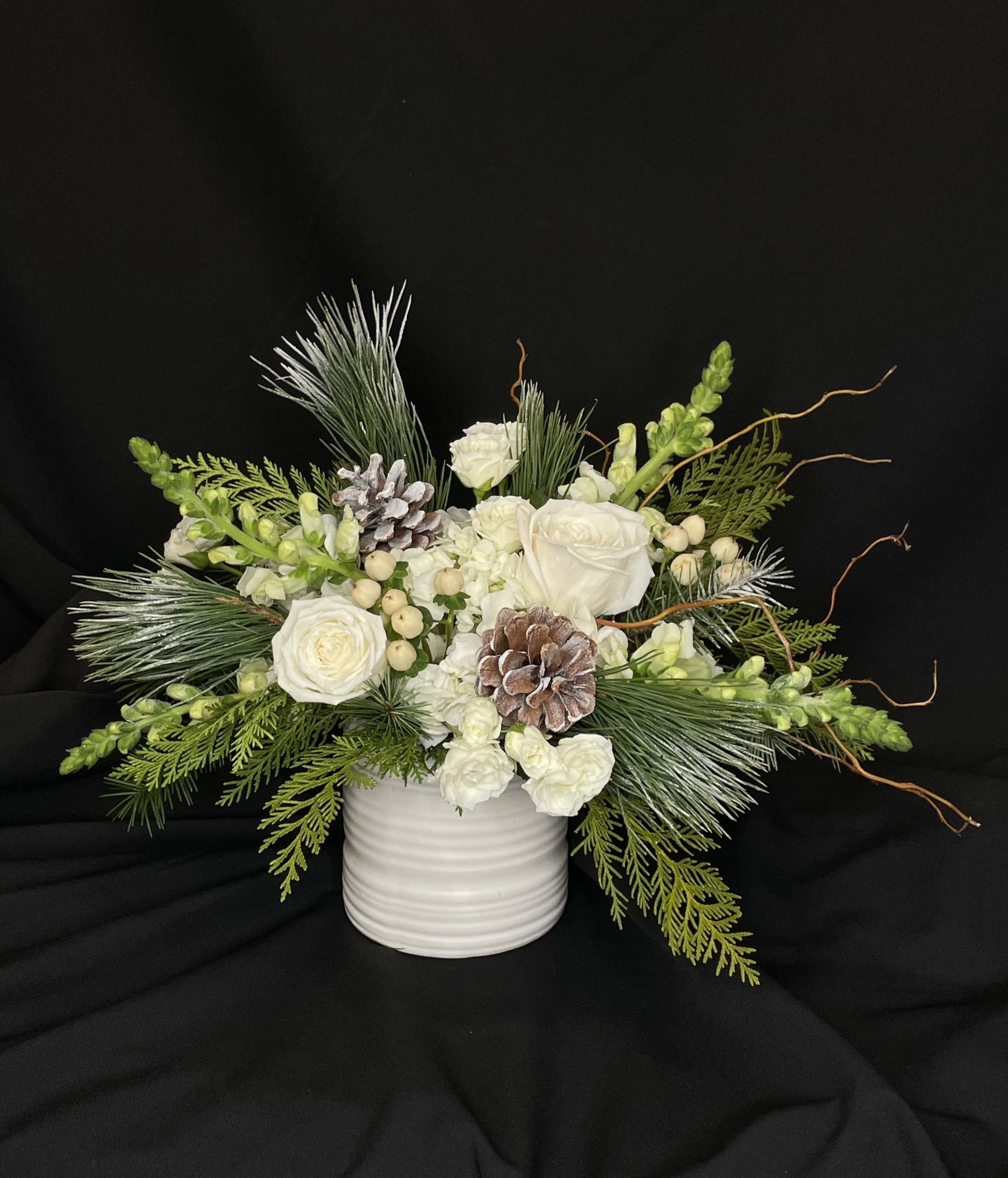 Forest Floor - This all white arrangement is the perfect way to celebrate the beauty of the winter season. Featuring branches, pinecones, hydrangea, roses, spray roses, snapdragons and hypericum berries, it shines with simplicity in a white ceramic vase. Perfect for those who love the natural elegance of the snow in the woods.