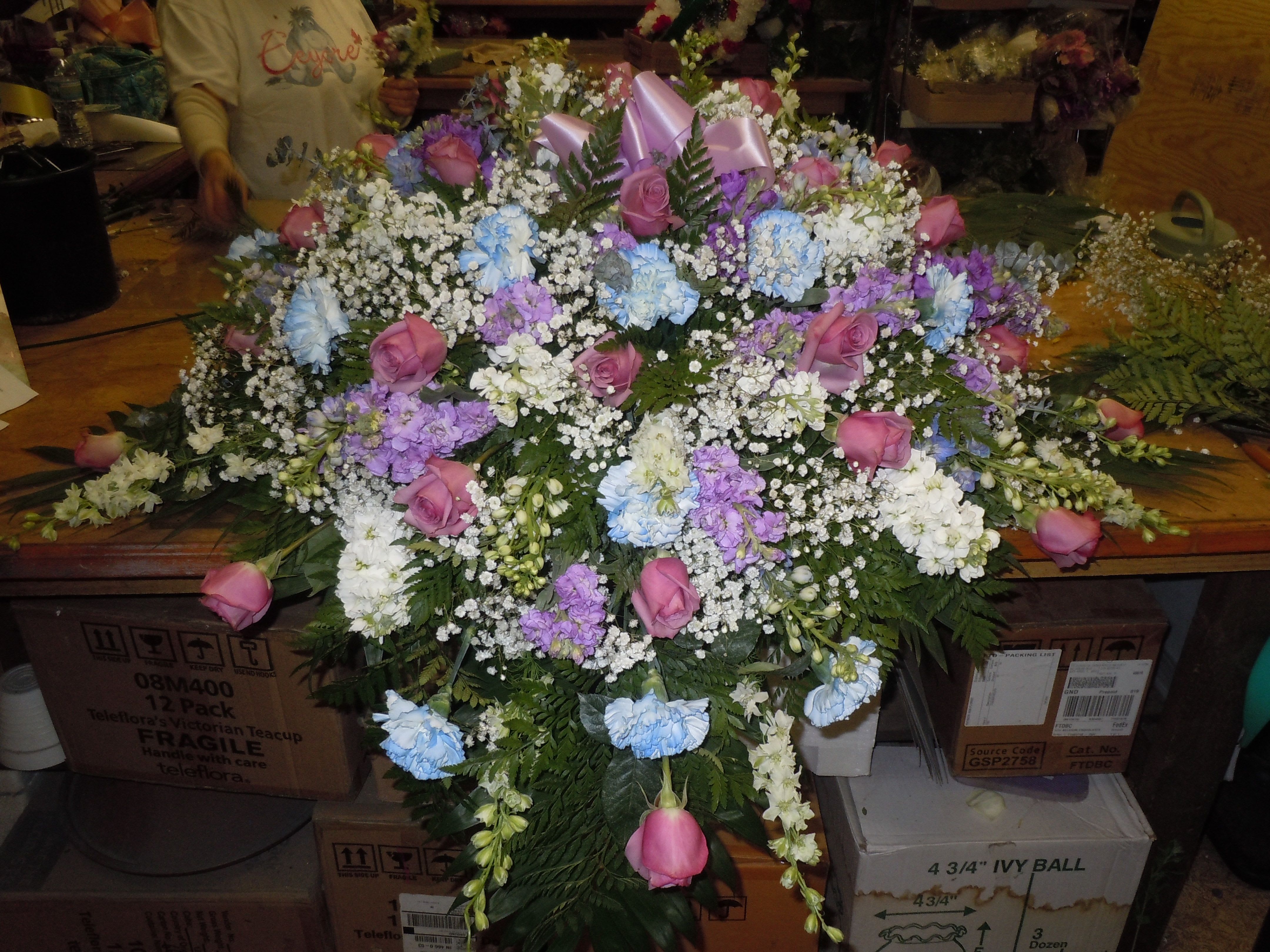 C25 Casket Spray - A mixture of white and lavender stock, lavender roses, white carnations tinted medium blue, whitesnap dragons, greens, and baby's breath, includes bow.  Banners are $15.00 + extra  Substitutions may apply, pending flower availability