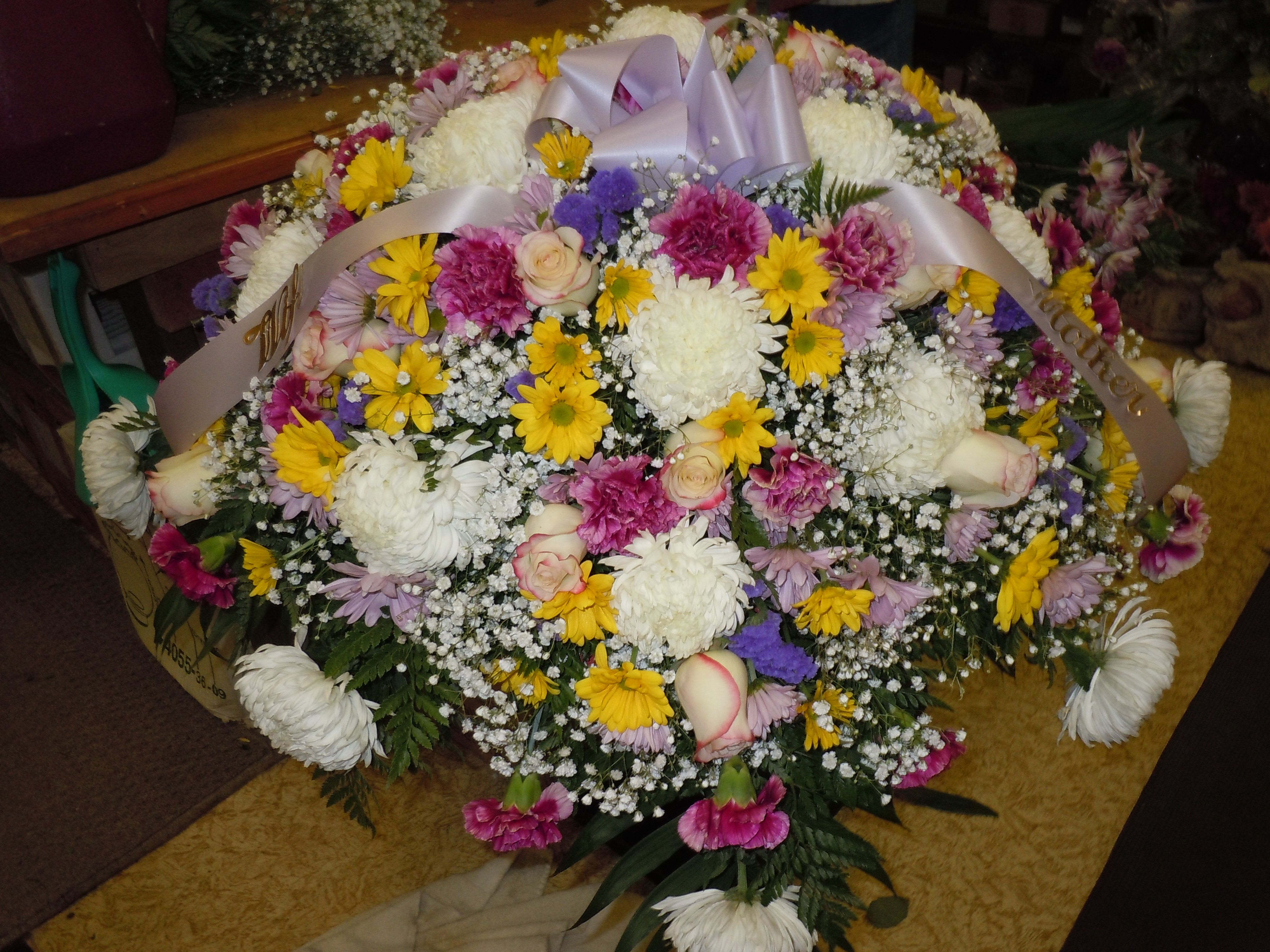C26 Casket Spray - A mixture of white fuji mums, light pink roses, orchid pink carnations, yellow daisies, purple statice, greens, and baby's breath, includes bow.  Banners are $15.00 +   Substitutions may apply, pending flower availability.