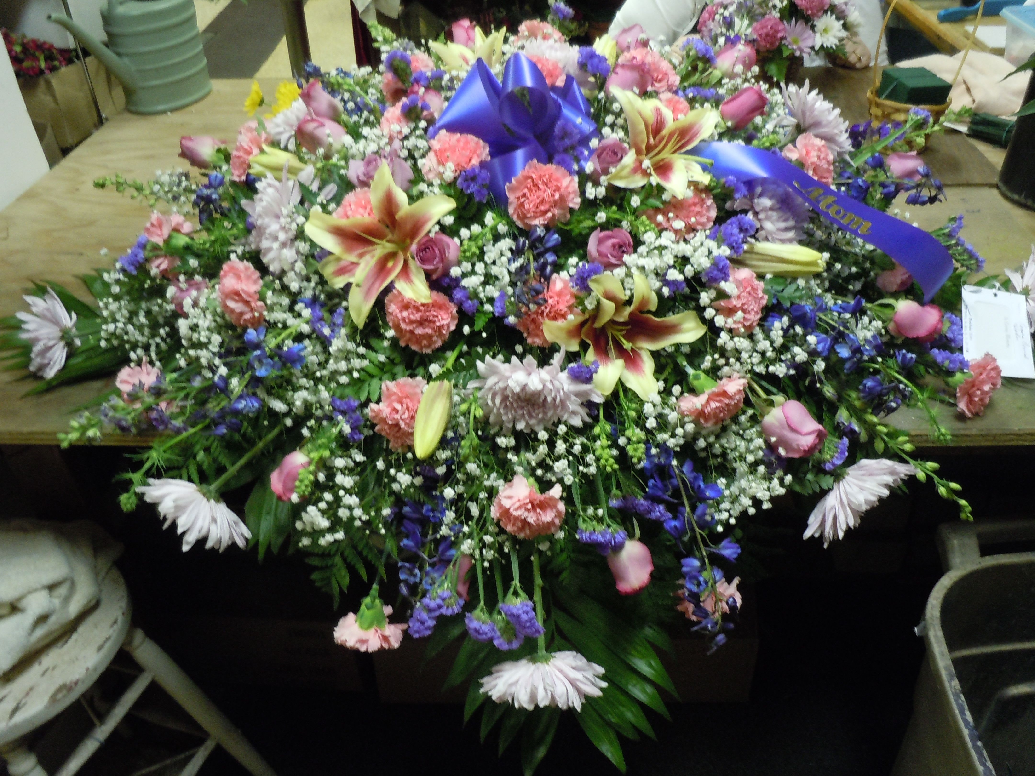 C29 Casket Spray - A mixture of lavender fuji mums, medium pink roses, pink carnations, blue delphinium. multi color lilies, purple statice, greens, and baby's breath, includes bow.  Banners are $15.00 +   Substitutions may apply, pending flower availability.