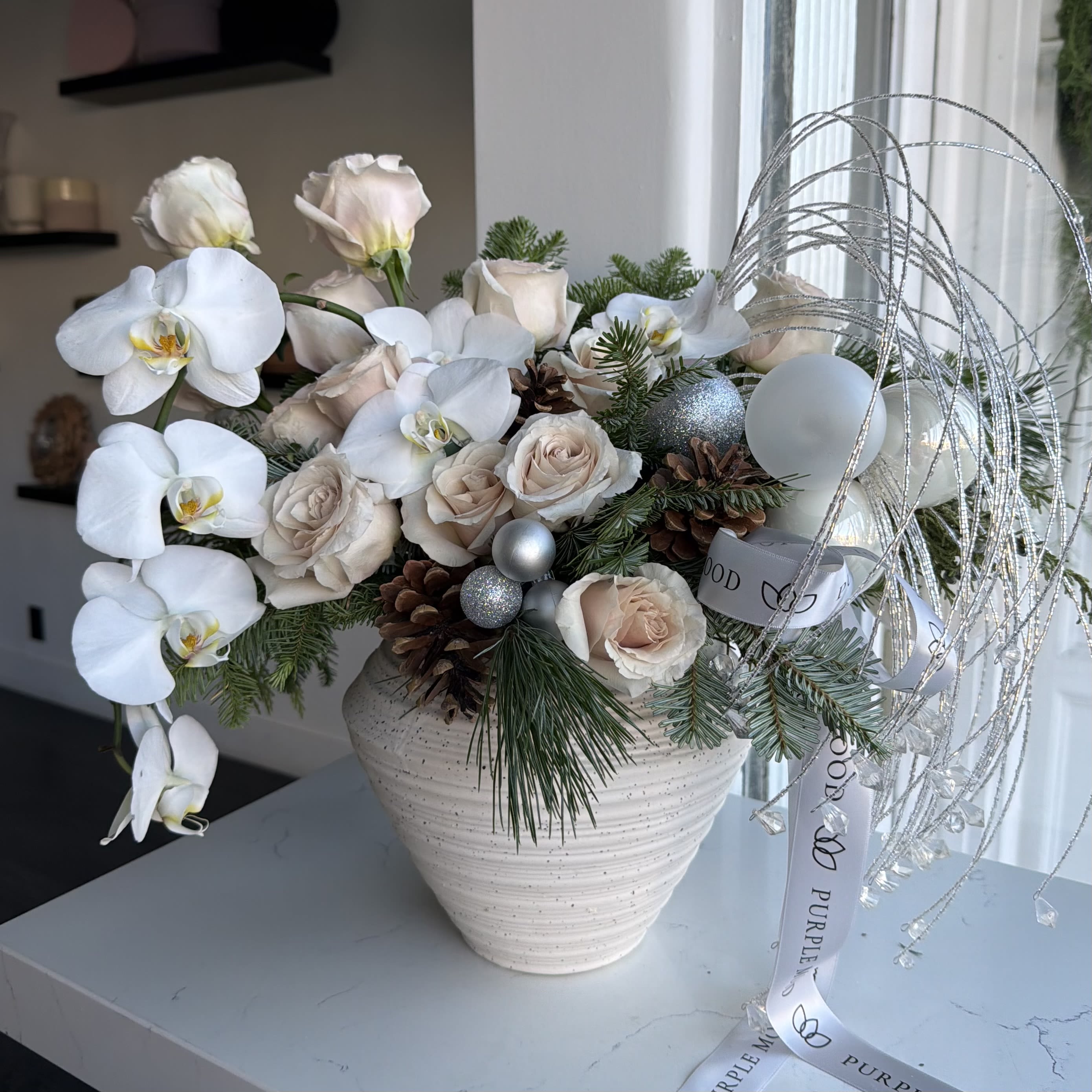 Winter Whisper Vase - A soft and elegant winter centerpiece designed to bring a touch of holiday magic into any space. Featuring cascading white orchids, premium blush roses, fragrant winter greens, pinecones, and shimmering silver accents, the Winter Whisper Vase blends modern luxury with cozy seasonal charm.  Finished with signature Purple Mood ribbons and arranged in a textured ceramic vase, this piece makes a stunning statement for holiday gatherings, gifting, or elevating your winter décor.  Perfect for homes, events, and corporate gifting. Same-day delivery available across Los Angeles. 