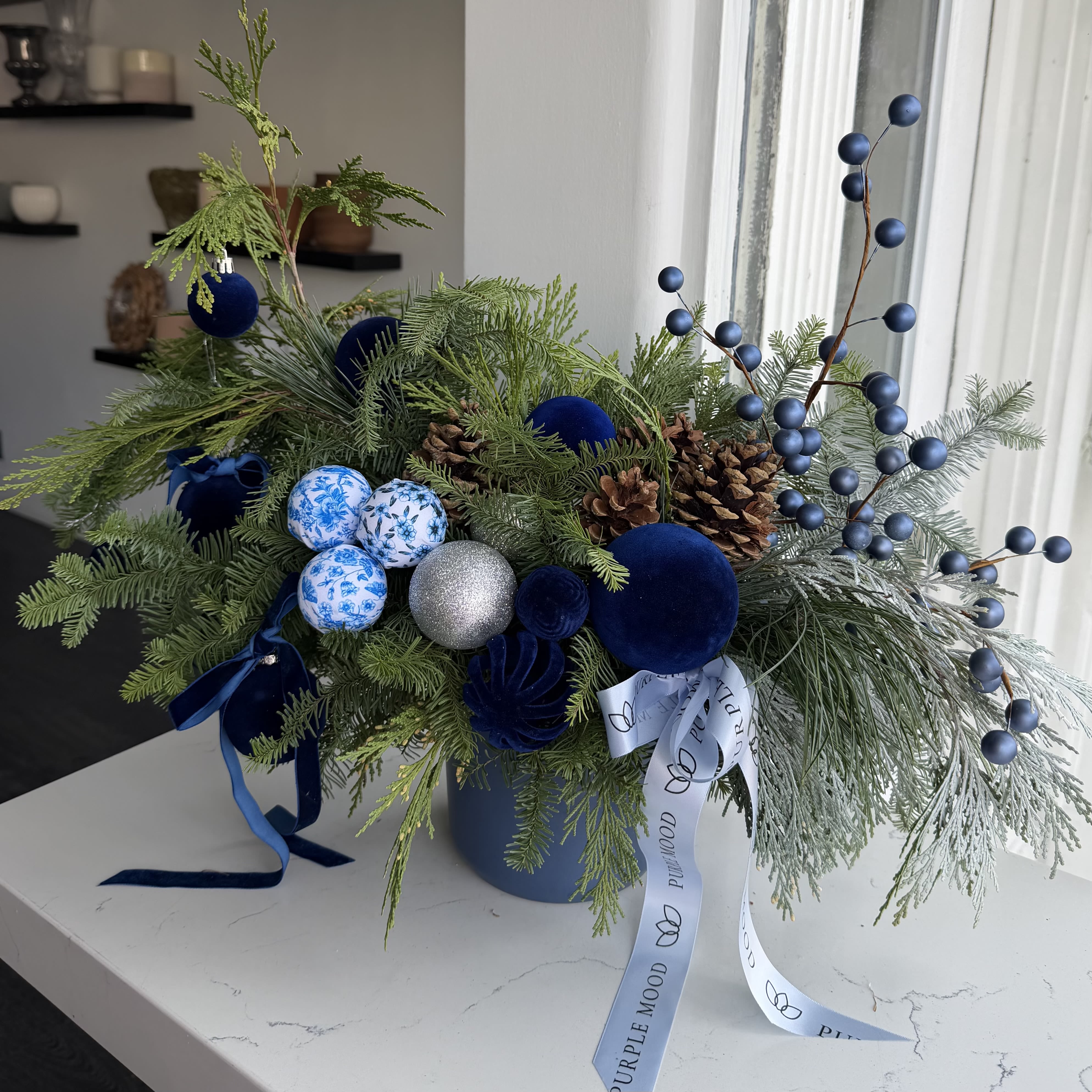 Blue Velvet Winter Vase - A luxurious winter arrangement created in deep midnight-blue tones with soft, velvety textures and elegant seasonal accents. The Blue Velvet Winter Vase features a lush mix of evergreens, pinecones, frosted foliage, velvet ornaments, matte blue berries, and delicate porcelain-style patterned spheres for a sophisticated, modern holiday look.  Arranged in a matching blue vase and finished with signature ribbons, this design brings a cool, refined winter elegance to any space. Perfect for gifting, holiday decor, or adding a unique touch of blue to your celebration.