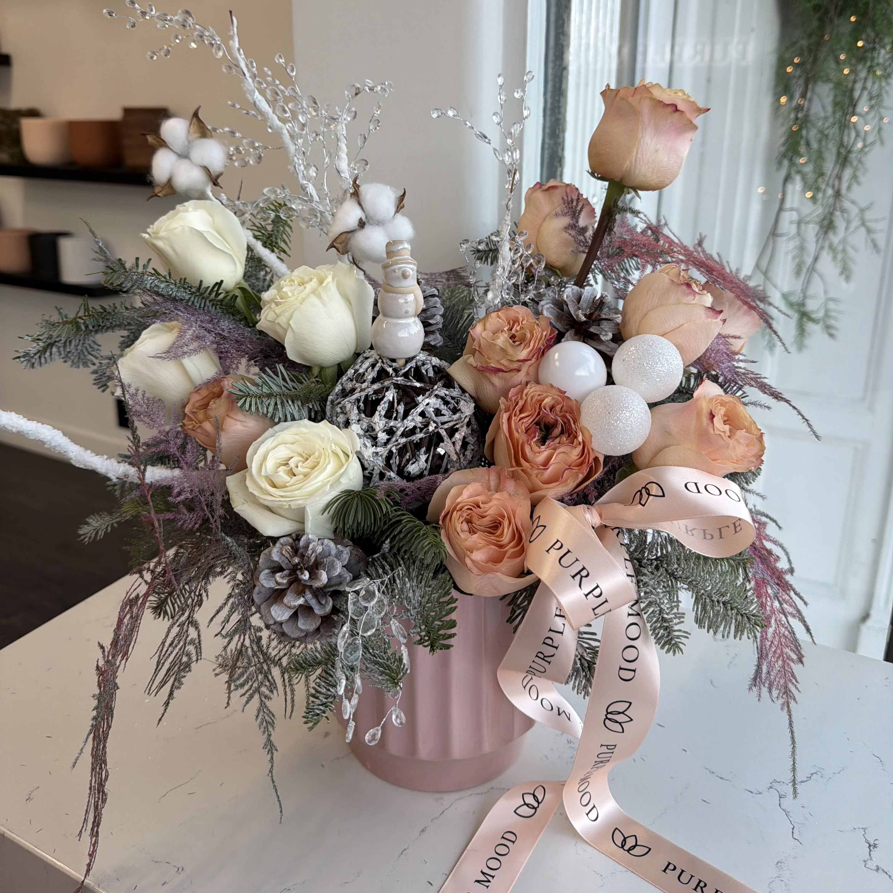 Peach Snowflake Vase - A delicate winter arrangement blending warm peach tones with crisp snowy accents. The Peach Snowflake Vase features premium peach and ivory roses, winter greens, frosted pinecones, glittering ornaments, cotton stems, and sparkling ice-crystal branches. A charming ceramic snowman adds a whimsical holiday touch, while the blush pink vase brings soft elegance to the entire design. Perfect for gifting, holiday table décor, or adding a cozy winter glow to your home. Same-day delivery available across Los Angeles. 