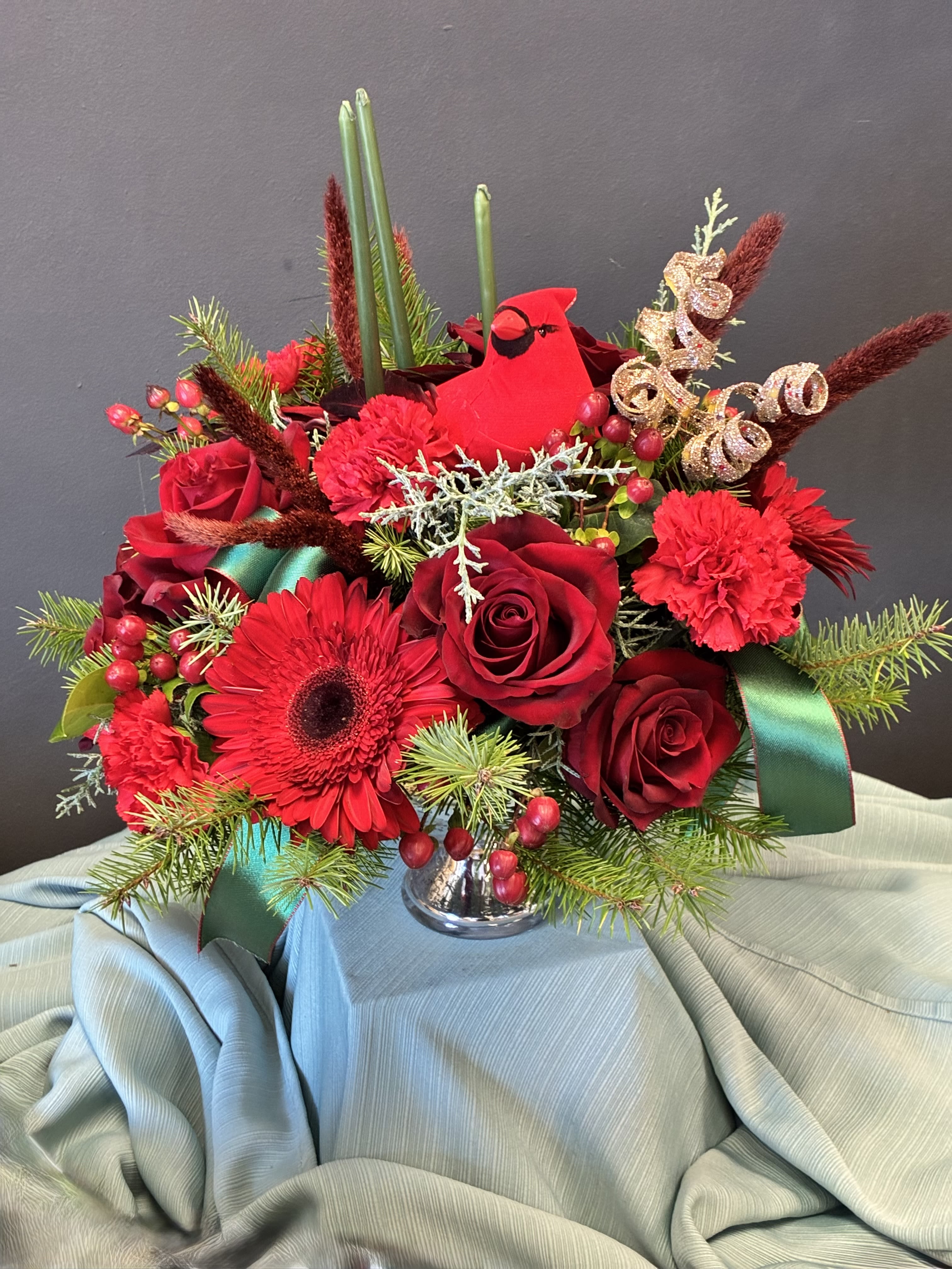  Cardinal Cheer Holiday Arrangement - This striking seasonal bouquet bursts with Christmas spirit, artfully arranged in a shimmering silver vase atop soft winter-blue fabric. Vibrant red roses, carnations, and a bold gerbera daisy evoke warmth and celebration, while pine branches and red berries bring the scent and texture of the holidays. A charming red cardinal perches among the blooms—symbolizing love, remembrance, and winter joy—accented by playful green ribbon and golden spirals that catch the light like tinsel. Tall green reeds and soft brown stems add graceful height, making this arrangement a perfect centerpiece for festive gatherings or a heartfelt gift to brighten someone’s season.