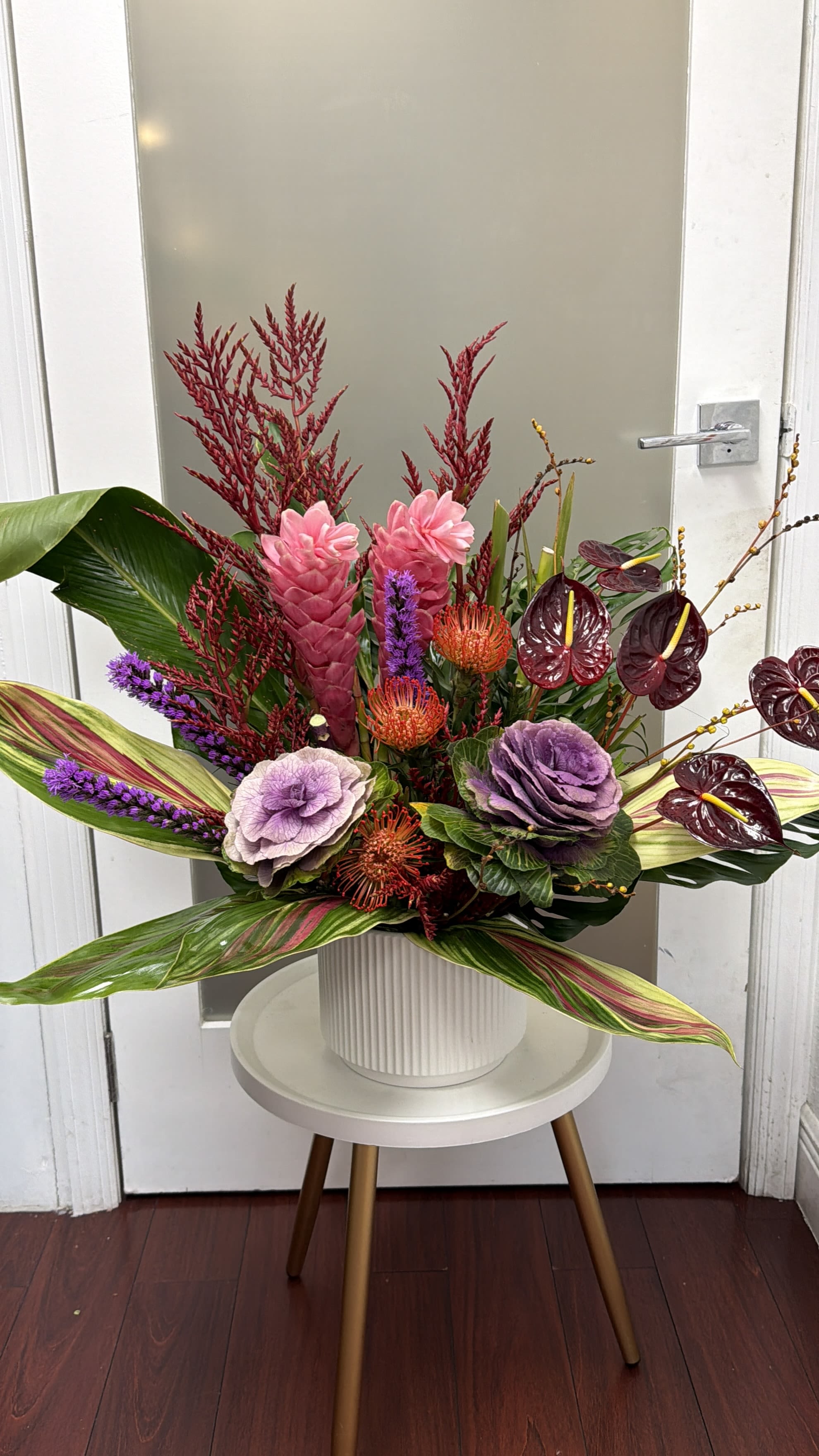 Coral Reef - A show-stopping tropical arrangement crafted in a modern white vase, blending bold colors and exotic textures. Pink gingers take center stage, surrounded by vibrant pincushion proteas, deep burgundy anthuriums, and elegant lavender accents. Ornamental kale adds depth and sophistication, while lush tropical foliage frames the design with dynamic movement and natural rhythm.  A captivating botanical piece perfect for upscale events, lobby décor, or making a dramatic statement in any space—radiating creativity, luxury, and tropical elegance.