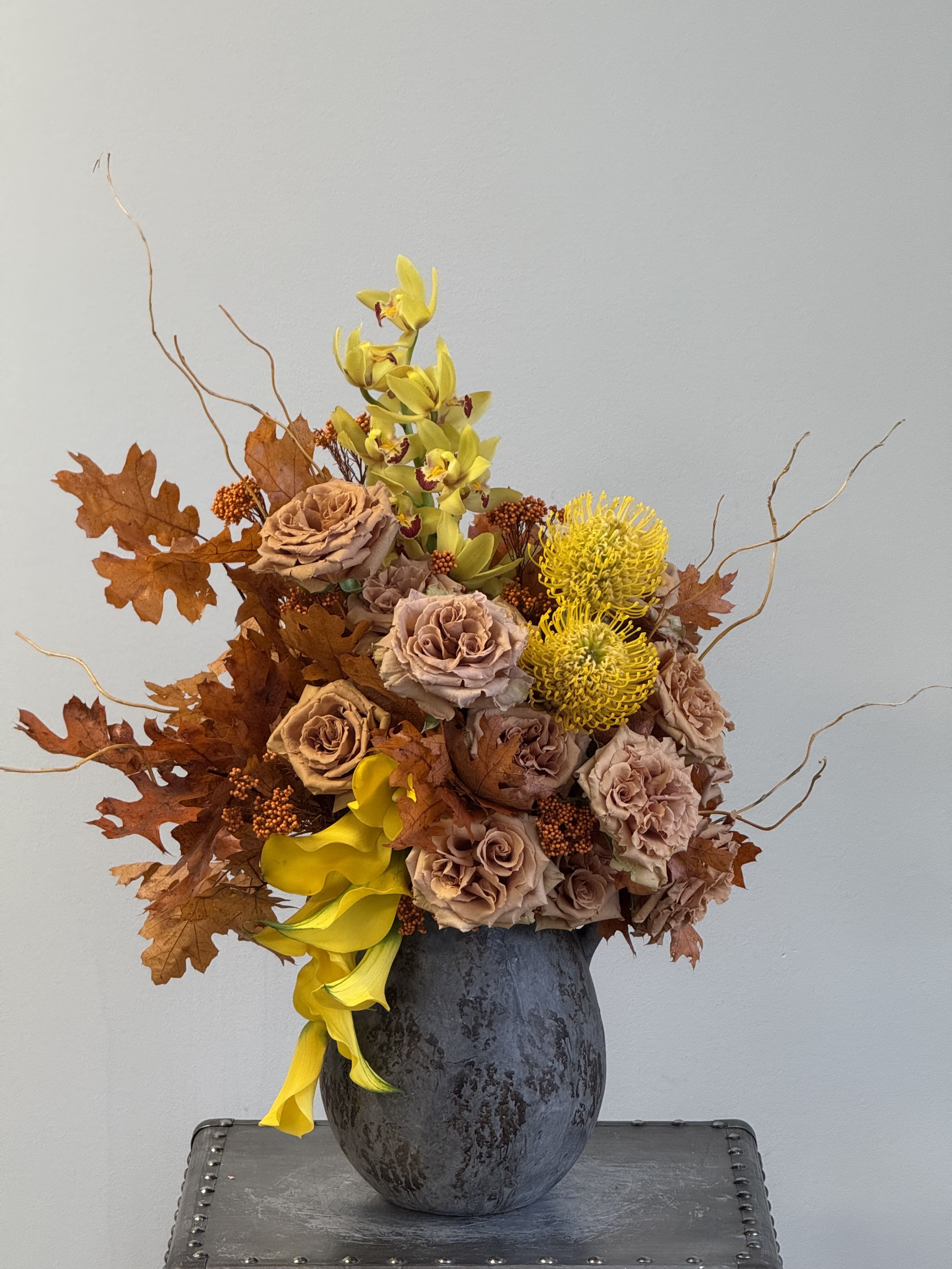 Golden Autumn Beauty - A striking arrangement of caramel roses, yellow orchids, and textural autumn foliage in a dark vas