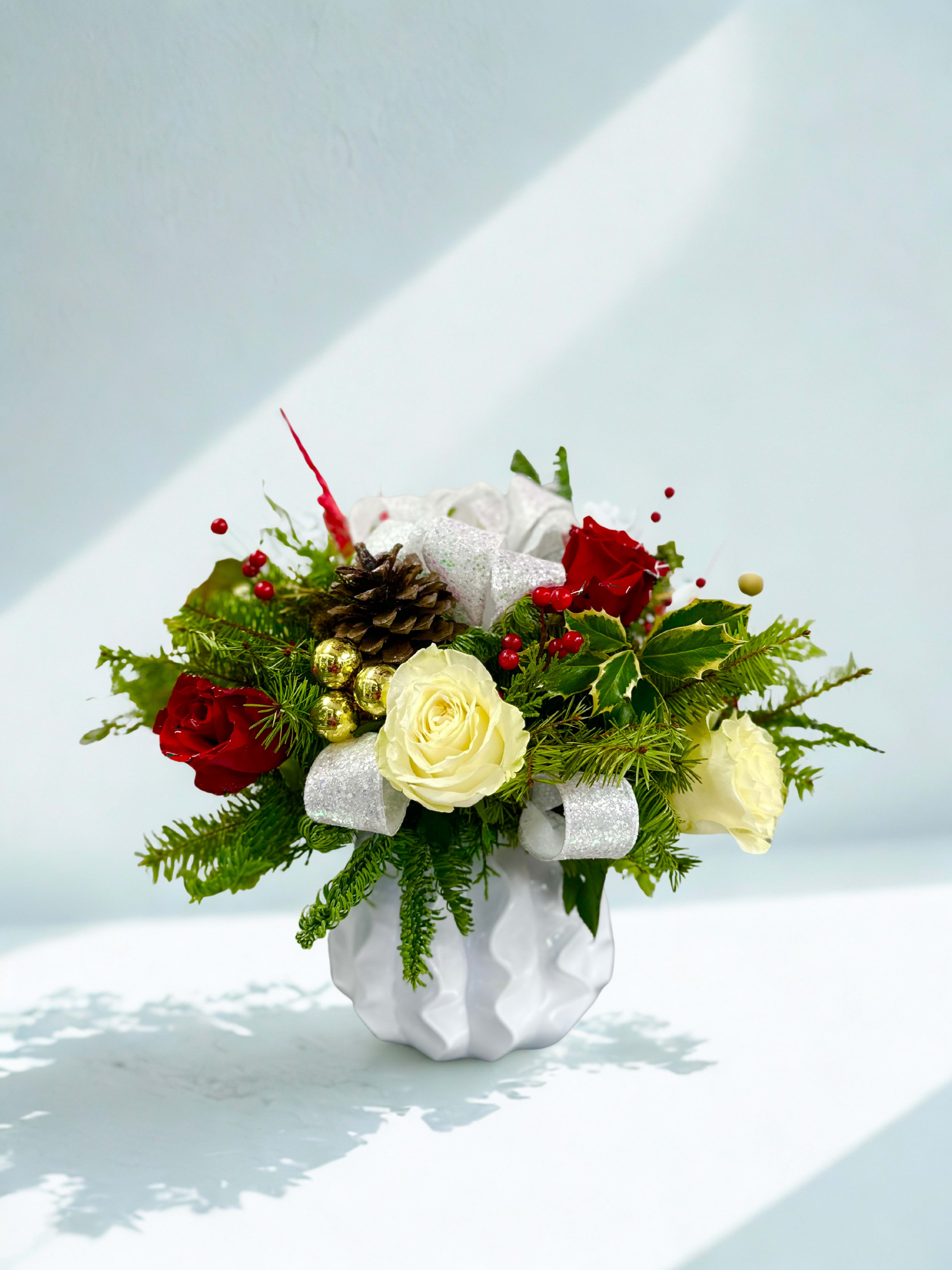 Winter Wonderment Arrangement  - Winter Wonderment is a festive and luxurious floral arrangement that captures the magic of the holiday season. Lush red and white roses are artfully arranged with a cheerful Christmas mix of seasonal evergreens, shimmering ornaments, and vibrant red berries. Sprigs of classic holly berries and a decorative holiday ribbon add rich texture and charm, enhancing the design’s warm, celebratory feel. Presented in a chic white designer vase, this arrangement blends elegance and holiday spirit, making it a stunning centerpiece for winter gatherings or a heartfelt seasonal gift. 