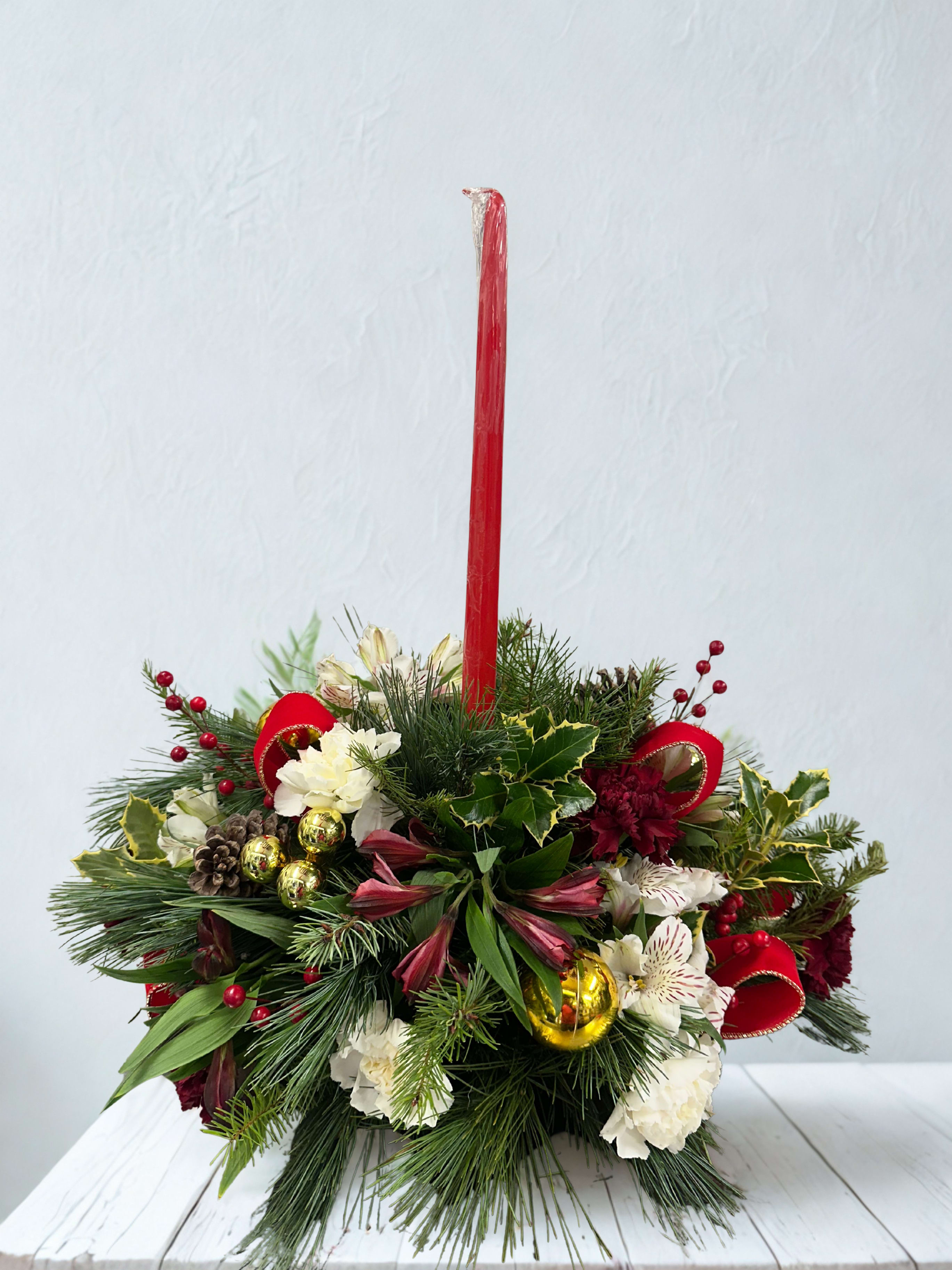 Holiday Cheer  - The Holiday cheer centerpiece features a lush blend of evergreen pine, holly, and seasonal foliage, beautifully accented with white and deep red blooms. Bright clusters of red berries, natural pinecones, and shimmering gold ornaments add warmth and cheerful sparkle. A bold red taper candle rises from the center, creating a classic Christmas touch that enhances the arrangement’s cozy, elegant charm. Perfect for holiday gatherings or as a striking seasonal display, this arrangement brings timeless winter beauty to any table