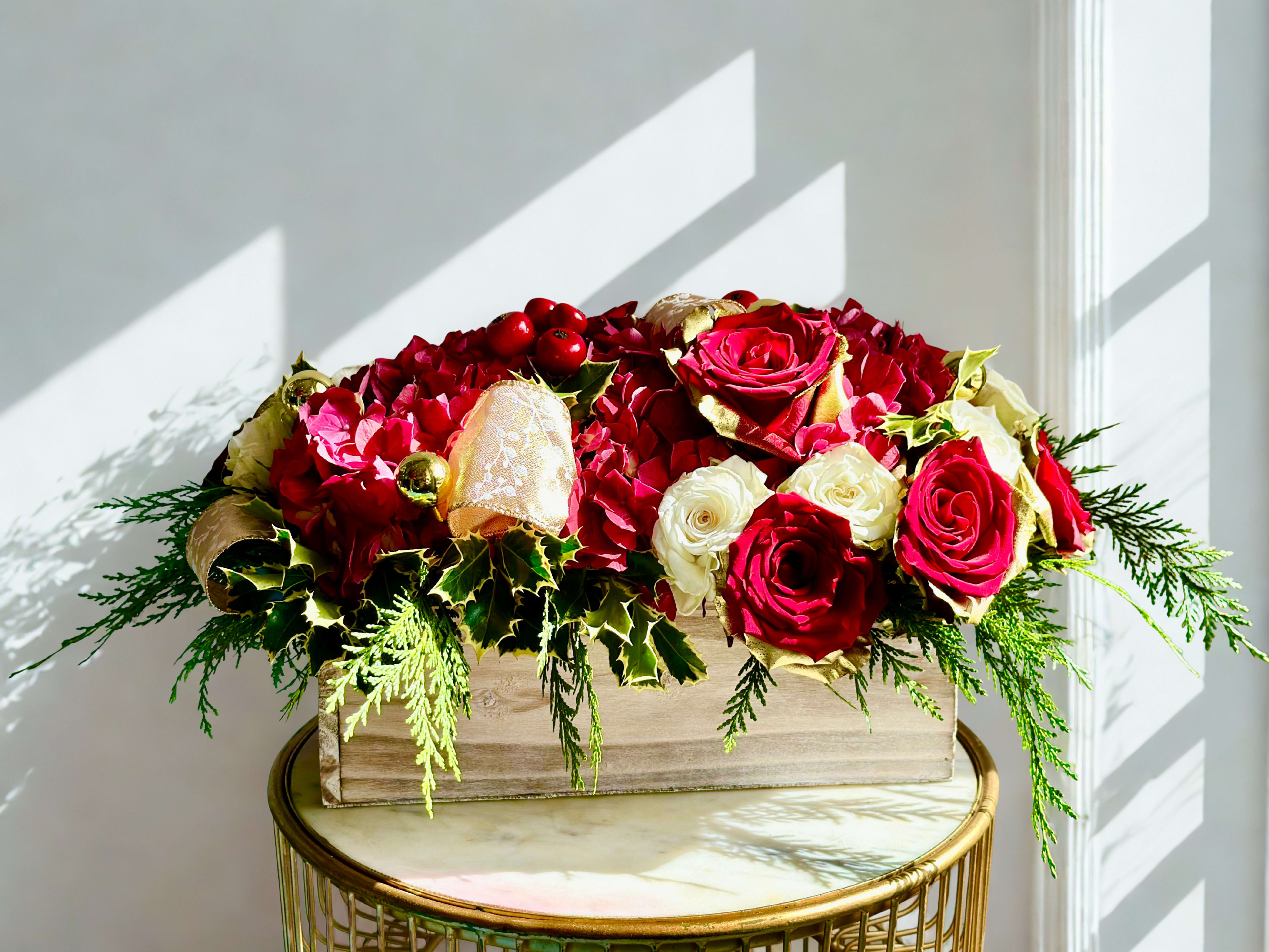 Cozy Winter Charm Floral Arrangement - The cozy Charm floral arrangement features a luxurious blend of red and white roses paired with vibrant red hydrangeas, creating a rich and festive color palette. Accents of holly, evergreen sprigs, and clusters of bright red berries add natural seasonal texture. Soft, shimmering touches of gold ornaments and gold-trimmed ribbon bring a hint of holiday sparkle. Designed in a rustic wooden box, this arrangement combines timeless elegance with cozy winter charm, making it a perfect centerpiece for festive gatherings or holiday décor.