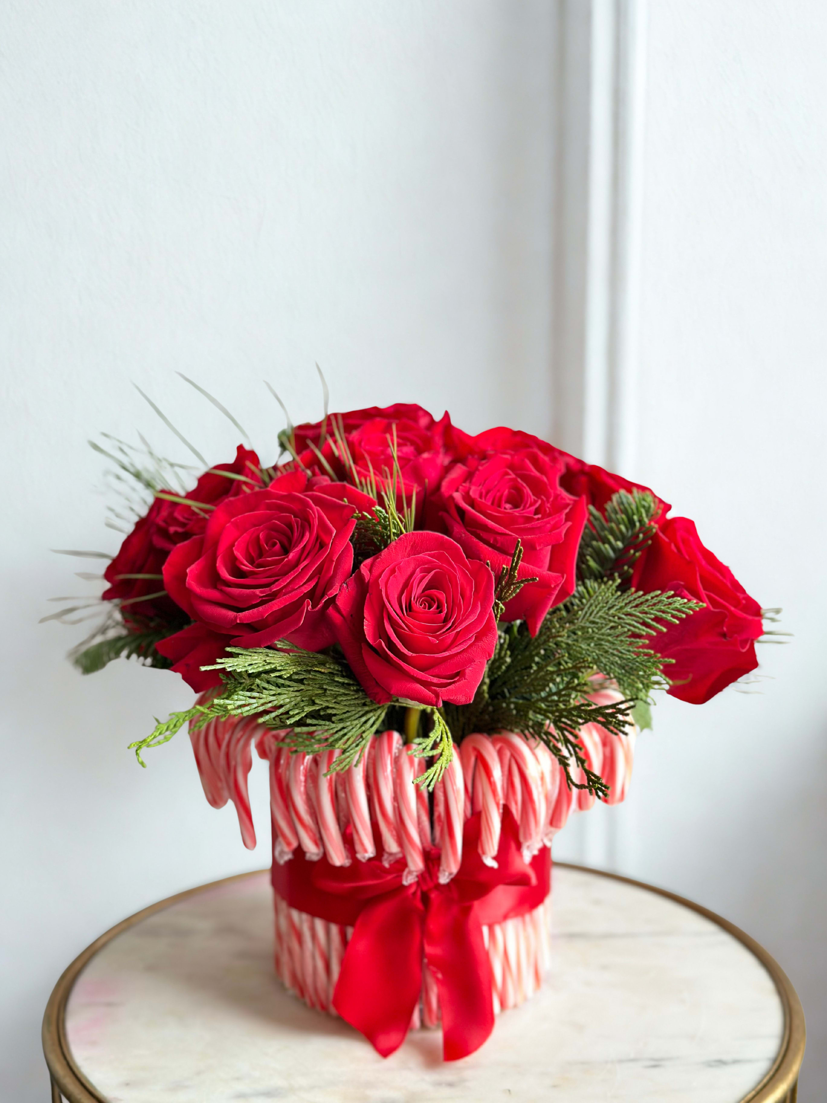 Welcome To Candy Land Arrangement - The Welcome Candy Land Arrangement is a playful and festive holiday display featuring radiant red roses nestled within a lush mix of Christmas pine. The rich greens and velvety blooms create a warm, enchanting contrast that captures the joy of the season. Presented in a whimsical candy cane–themed vase, this arrangement brings a touch of sweet holiday magic to any space—perfect for welcoming guests or adding a cheerful, nostalgic charm to your décor.