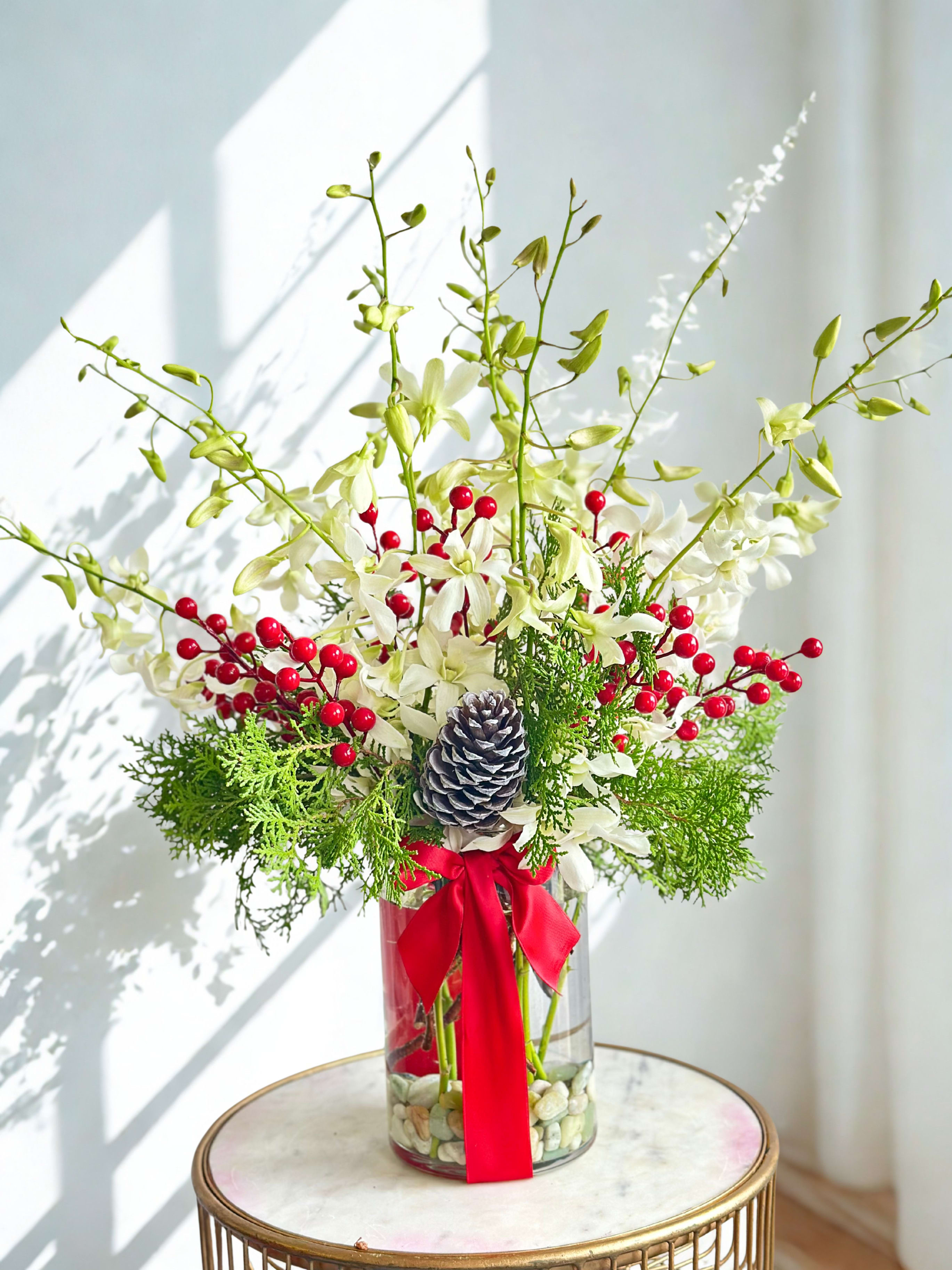 Let It Snow Floral Arrangement  - Let it snow floral arrangement featuring elegant white orchids nestled among fragrant fresh pine branches. Accents of vibrant red berries add a festive pop of color, creating a striking contrast against the soft orchid petals and deep green foliage. The combination of graceful blooms, natural evergreens, and seasonal berries brings a sense of refinement and holiday warmth, perfect for elevating any space with a touch of serene, wintry beauty. 