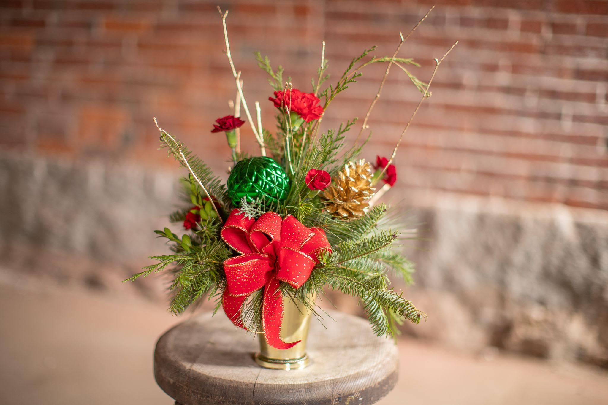 Happy Host - A perfect festive sentiment for your host, teacher,  friend, neighbor or co worker. A gold or silver julip container is carefully filled with fresh inter foliage varieties. Incense, Port Offord cedars, balsam, pine and Carolina Sapphire bring the  season's essence to life. Decorative pine cones in either silver or gold to match the container are carefully selected with coordinating branch materials. A seasonal ribbon will be selected for a festive handcrafted bow to complete this simple designs. Standard size will include carnations and mini carnations for floral selections. Upgrading to deluxe will include roses. This harmonious arrangement is perfect for merry-making this seasons!