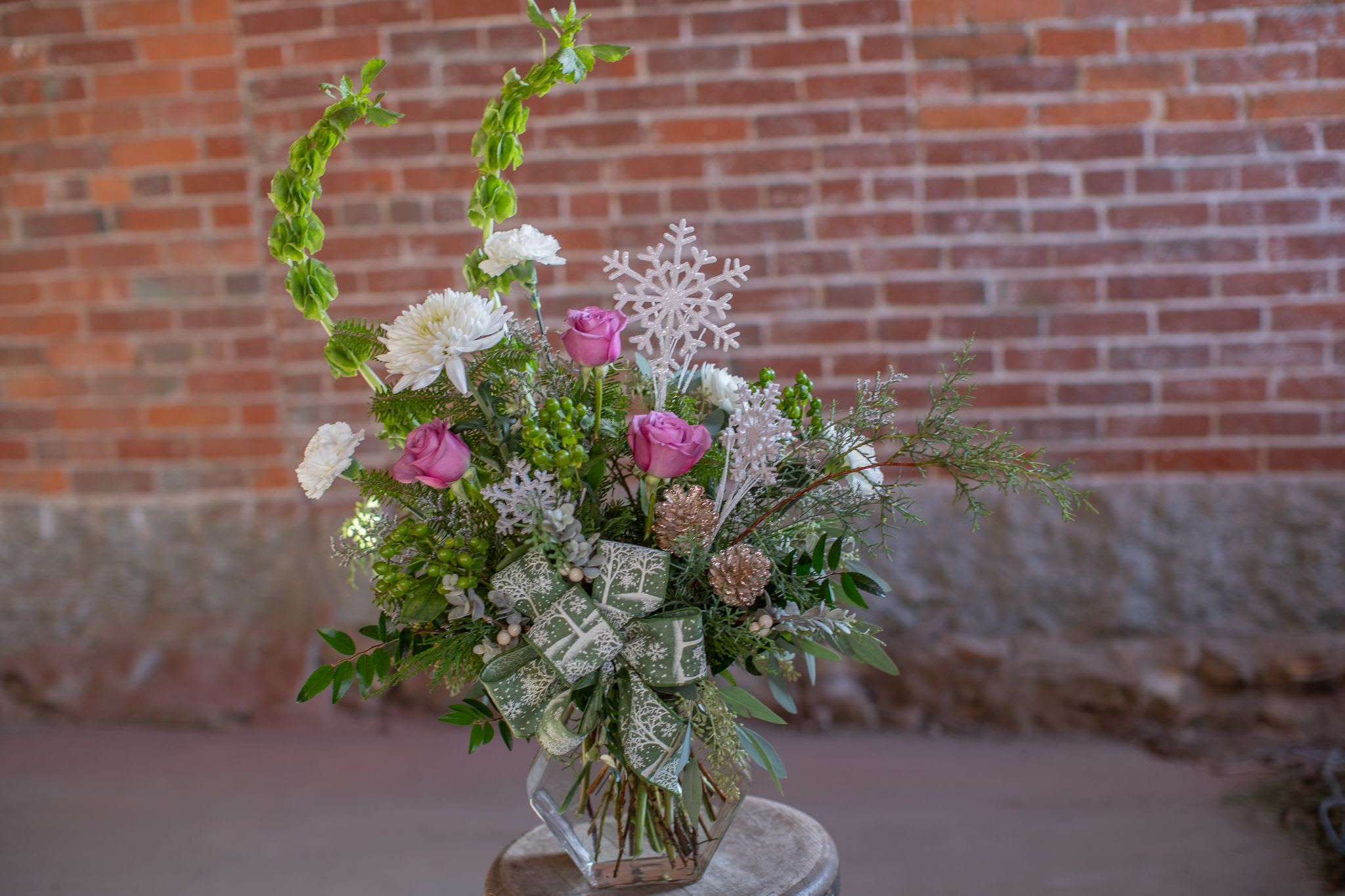 Hexagon Flurry - This one sided design is featuring festive green bels of Ireland, hypericum berry, white dianthus and cremons. Mauve toned roses are photographed but may be more lavender based on our rotating inventory. Decorative glimmering snowflake pics are carefully placed above the flowers in this vase design. Vase is a hexagonal geometric style glass jar just as snowflakes are sacred ice geometry. A more colorful spin for the season instead of traditional red and white. Winter scene printed ribbon completes the vase of flowers. 