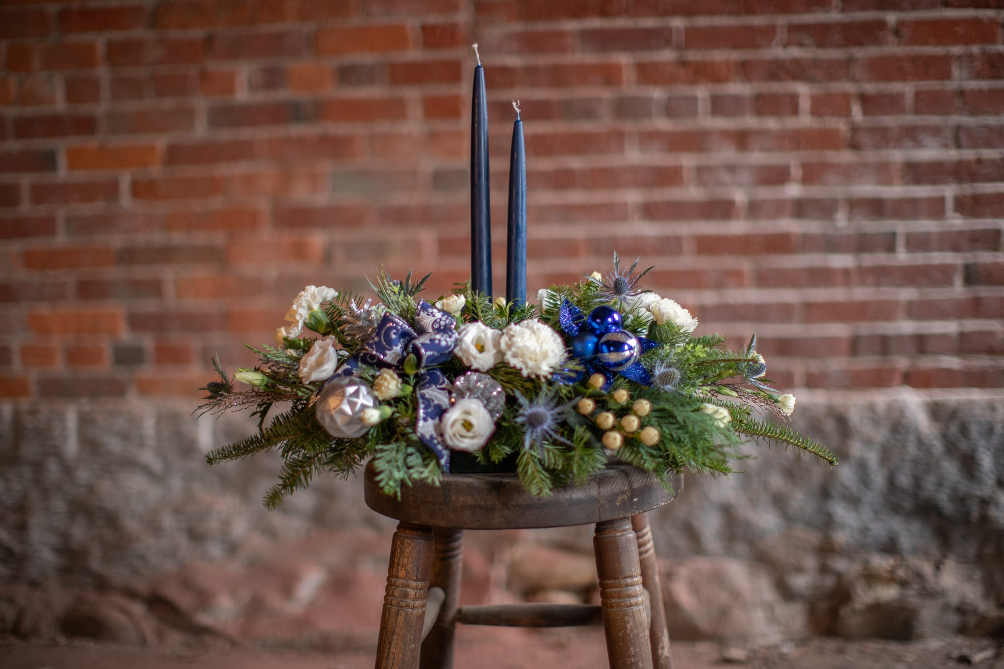 Indigo Icicles Centerpiece  - Winter foliage with white and cream flowers are a perfect pair with indigo candles, silver decorative items, blue thistle and navy ribbons. Staggered slow burning taper candles light up the night and sparkling silver pine cones add warmth to this cool, icy design. Traditional long and low centerpieces are a timeless holiday classic. Add to the festivities and your beautiful place settings as you entertain your friends and family alike. 