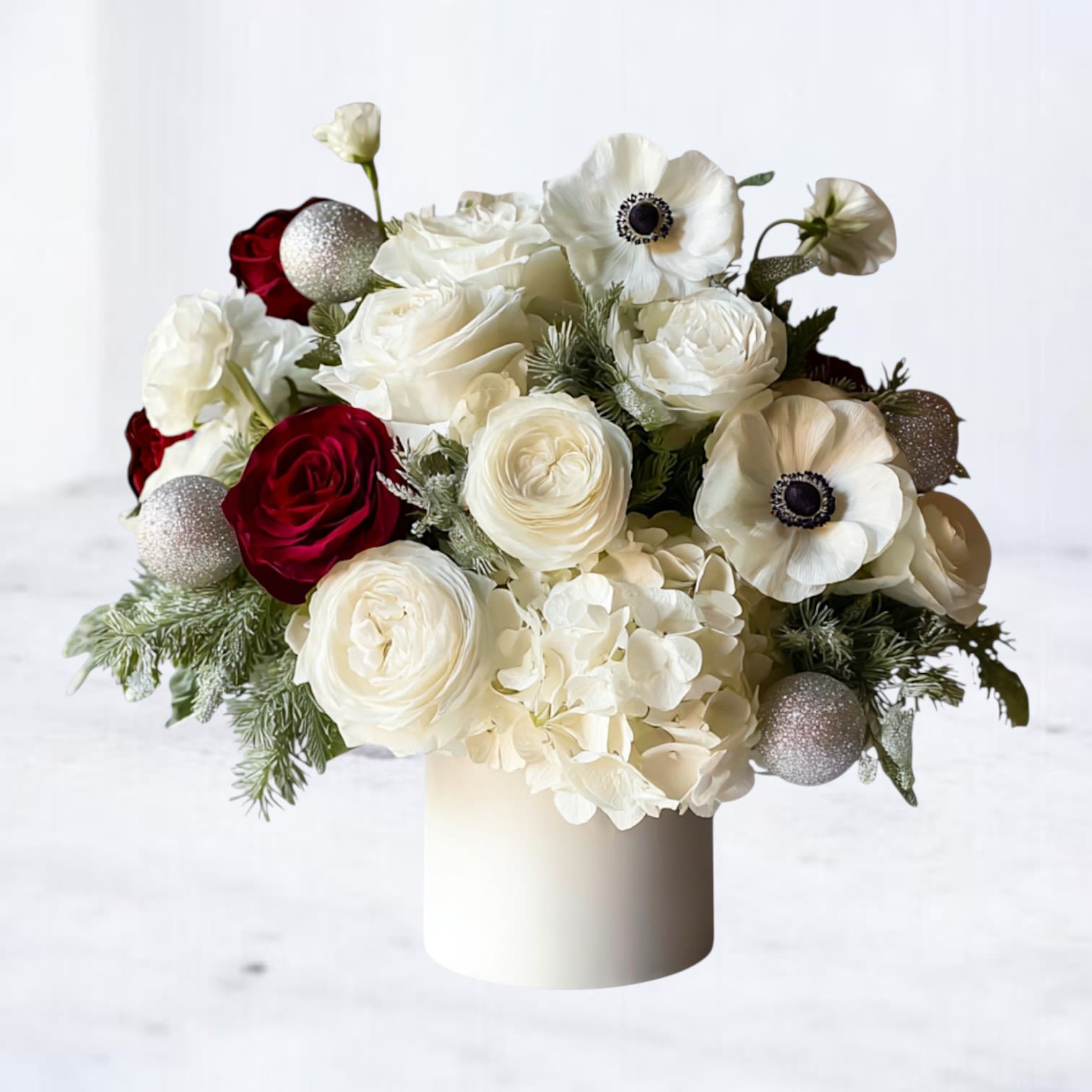 Nordic Winter - A modern winter statement piece inspired by Scandinavian minimalism and luxury holiday styling. Nordic Winter blends crisp whites, frosted textures, and bold pops of deep red to create an elevated seasonal design that feels timeless, artistic, and effortlessly chic.  This centerpiece features premium white roses, hydrangea, anemones, and ranunculus paired with lush winter greenery, icy accents, and sparkling ornament details. Rich red roses add a striking contrast, giving the piece depth and warmth while maintaining its clean winter aesthetic. Designed in a modern white cylinder vase, it brings high-end holiday elegance to any space—perfect for coffee tables, entryways, dining tables, and gifting.  Whether you're hosting for the holidays or sending a truly memorable seasonal arrangement, Nordic Winter delivers a polished luxe look that stands out from traditional holiday flowers.