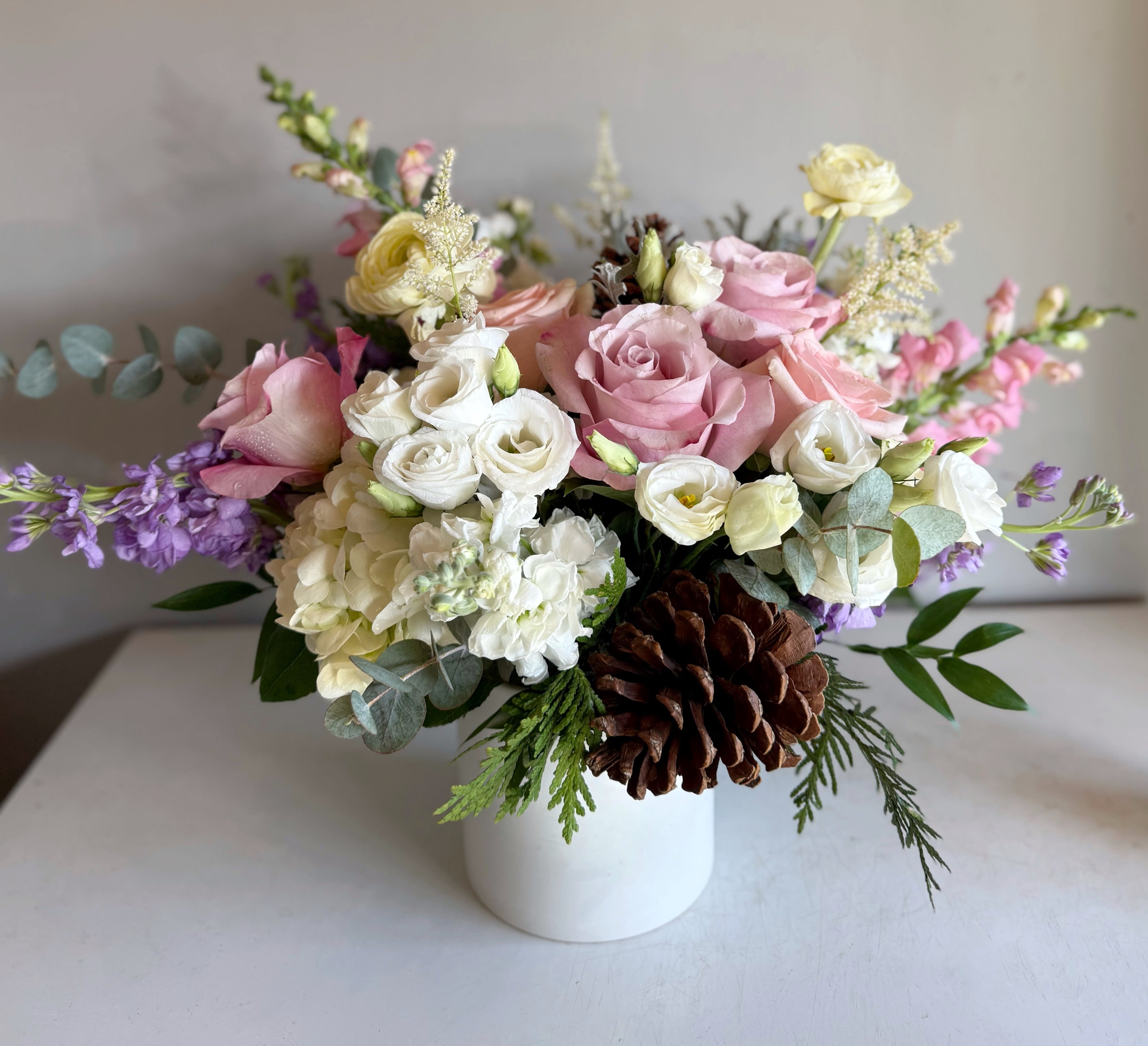 Winter Blush - With blush petals, frosty white accents, and a hint of lavender glow, this arrangement brings a touch of winter magic in a tender, pastel palette. Featured flowers include Lisianthus, Secret Garden roses, Snapdragon, Stock, hydrangeas, and winter evergreens.
