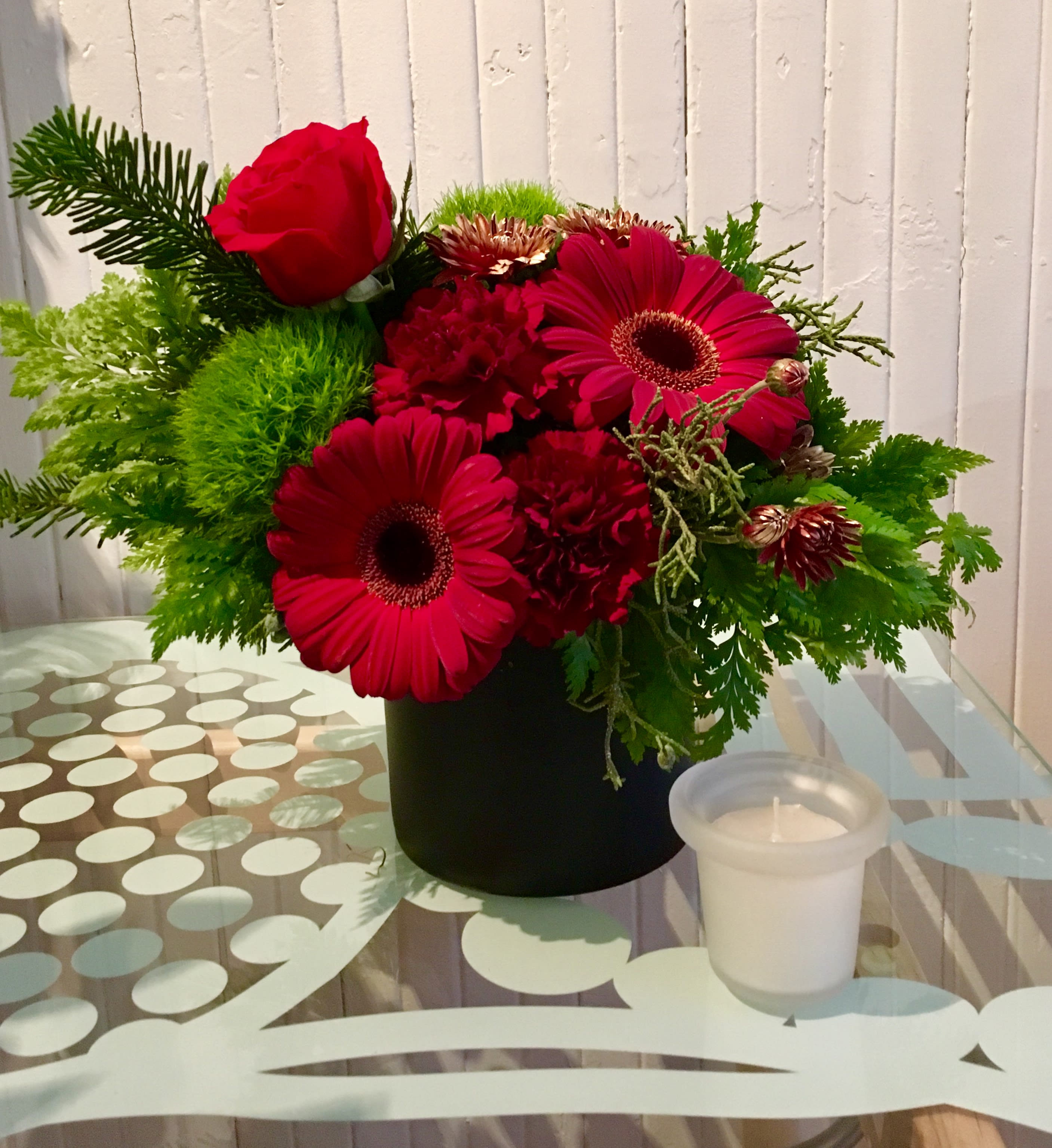 Happy Kwanzaa.  A Floral Archist™ original. - ‘Happy Kwanzaa’ The first fruit of our ‘Winter Celebration Collection’.  This beautiful custom designed floral piece is the perfect way to wish your loved ones a beginning of a blessed season. Gerberas, Roses, Dianthus and an aromatic pines collection of everygreens in a black container.  As with ALL our designs, selection of blooms vary with each Upgrade. As part of our Winter Celebration Collection, this design serves ALL winter celebrations.   NOTE: Our flowers and greens are a gift from nature. It’s shade and depth might vary, design, aesthetic and value remains the same. In support of our efforts to aide in the preservation of our planet, PJf uses Fair Trade flowers, Plants, and Recycled containers. Palette is seasonal.