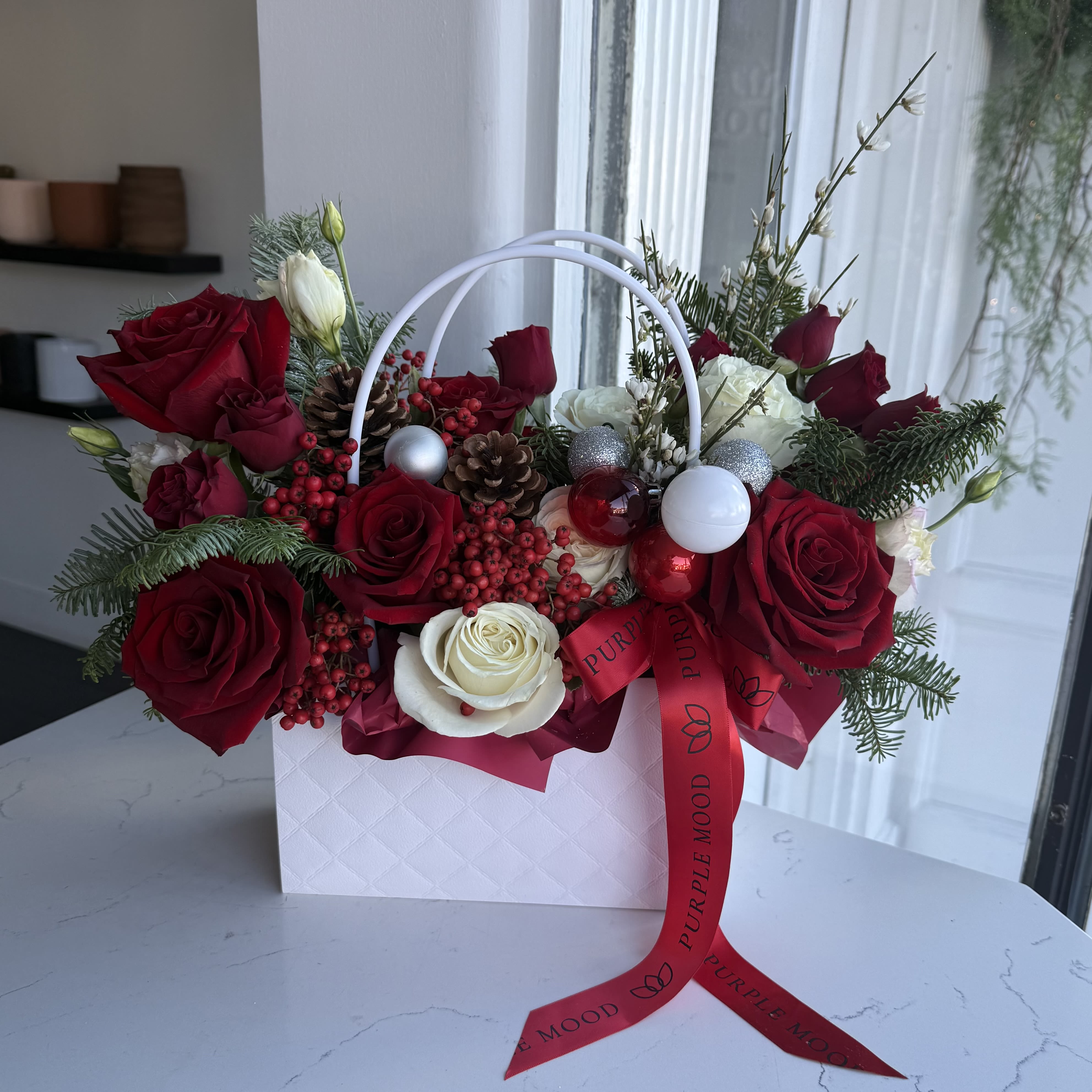Christmas mini Bag - A charming holiday arrangement designed in a white quilted mini bag with lush red roses, creamy blooms, festive berries, pinecones, and sparkling ornaments. Elegant, compact, and full of Christmas magic — perfect for gifting or decorating any space.