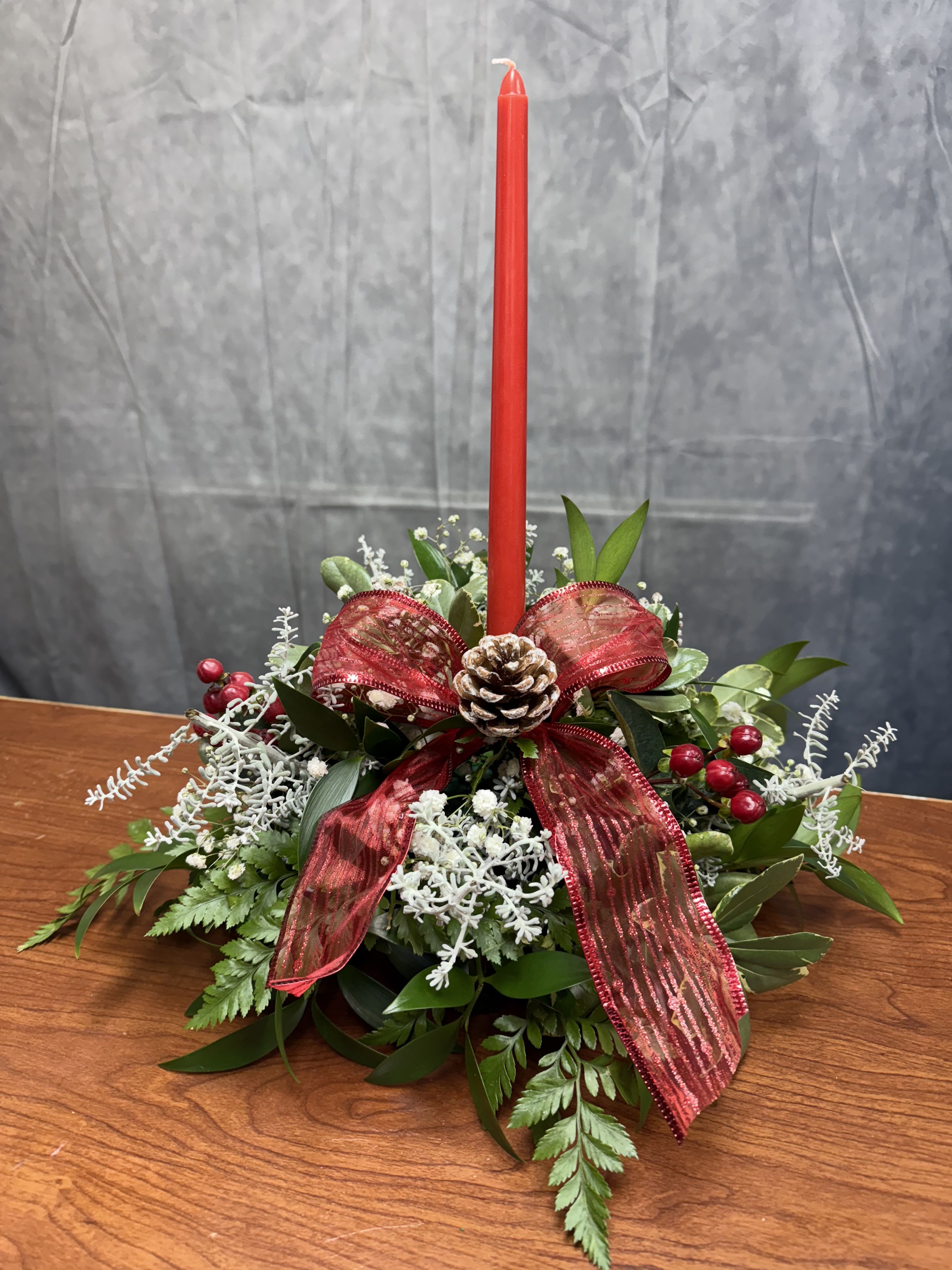 Pinecone Joy - Add a touch of holiday warmth with our 12-inch circular pinecone joy. Crafted with lush Christmas greenery, natural pinecones, vibrant red berries, a festive bow, and a red candle at its heart, this low-profile design is perfect for any table setting.  Candles colors can vary depending on the availability (red, gold , white)