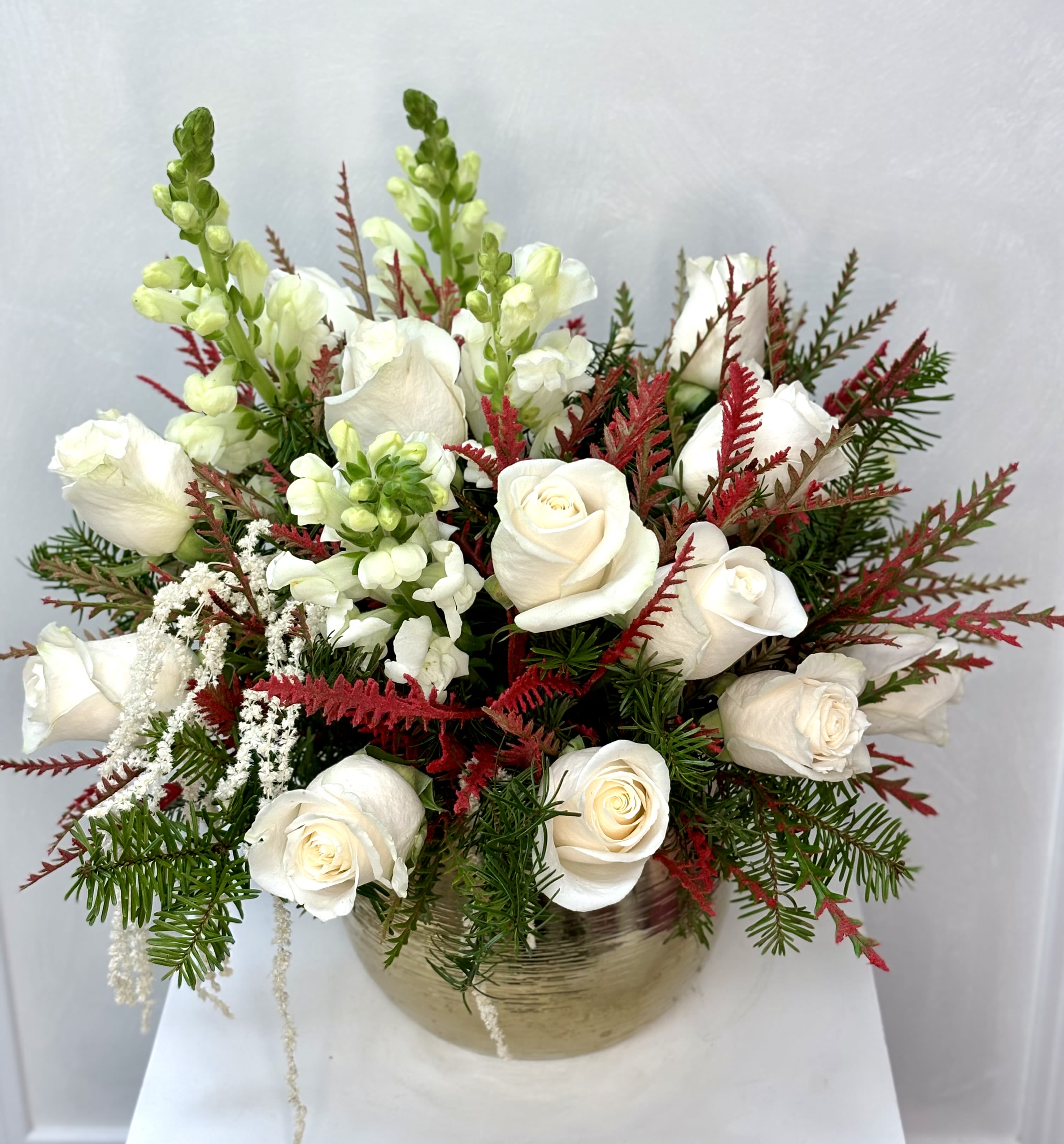 Winter Whisper Elegance By VLM Flowers - A graceful winter arrangement featuring creamy white roses and soft white snapdragons accented with cascading astilbe. Rich evergreens and feathery red foliage add warmth, contrast, and a festive seasonal touch. Designed in a stylish gold-toned vase, this piece brings serene winter beauty to any space—peaceful, refined, and timelessly elegant.